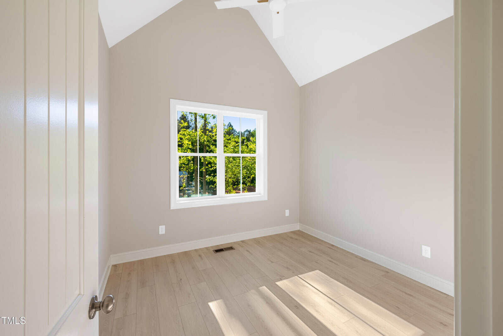 A small, empty room with light-colored wood flooring, beige walls, a white ceiling fan, and a large window letting in natural light with a view of green trees outside.