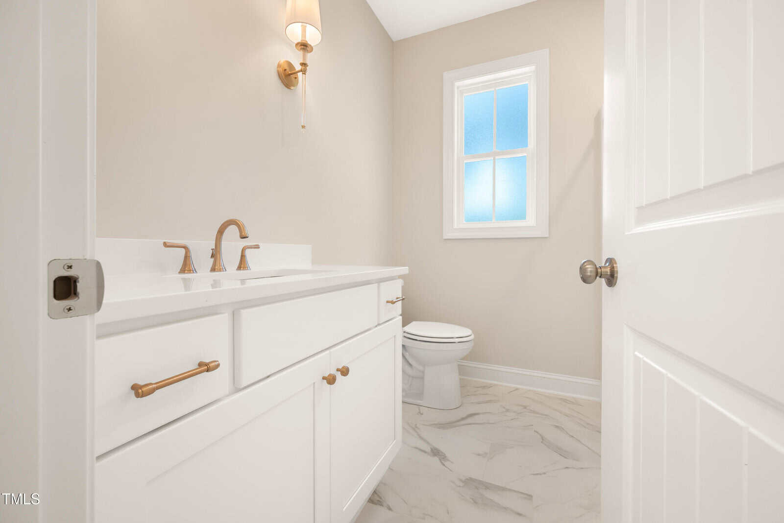 A bathroom with white cabinets, gold hardware, a white countertop, a single-mounted light fixture, a toilet, and a window letting in natural light. The floor has a white and gray marble pattern.