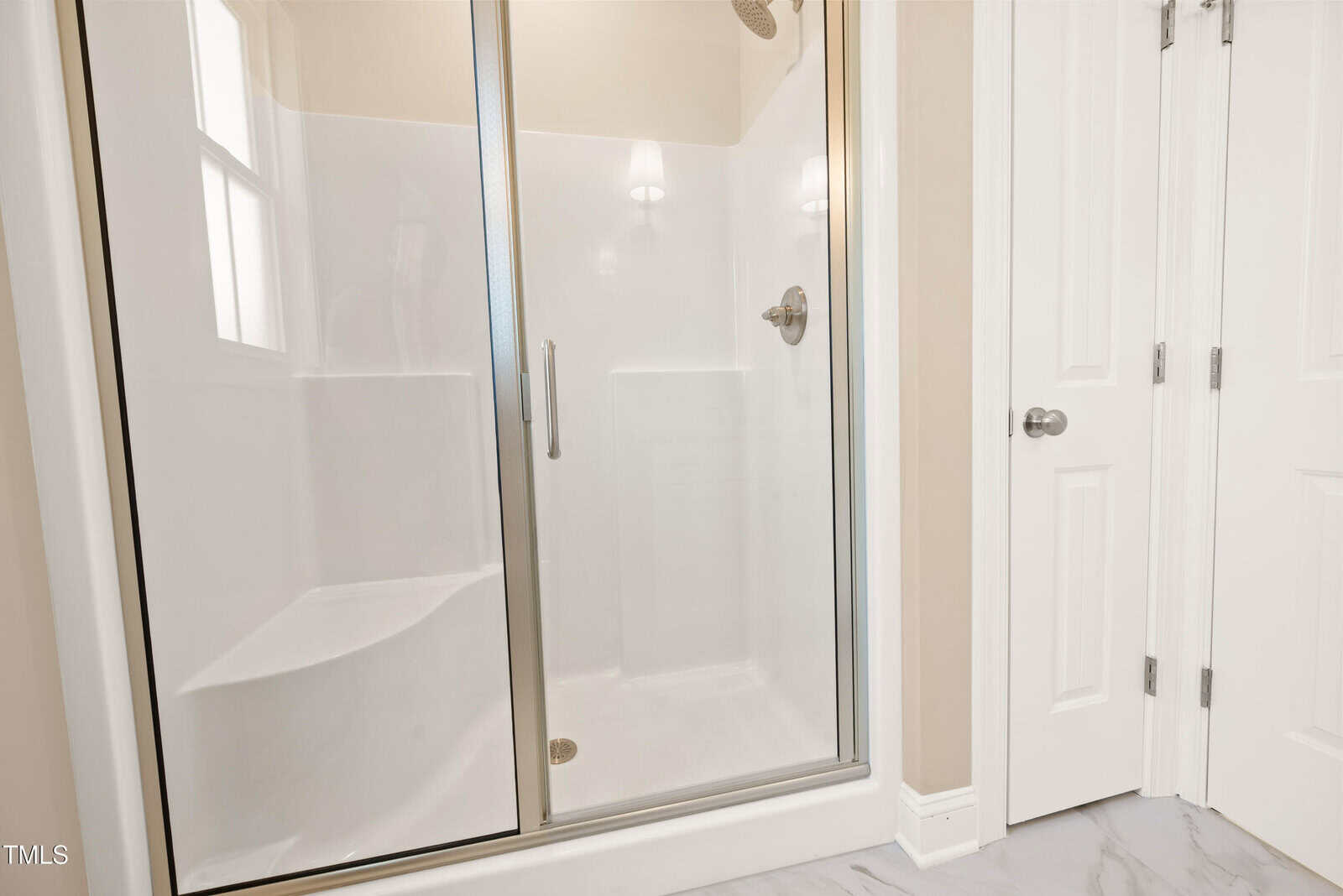 A bathroom with a glass-enclosed shower featuring a built-in bench and a metal showerhead, next to two white paneled doors and a window. The floor is finished with light-colored tiles.