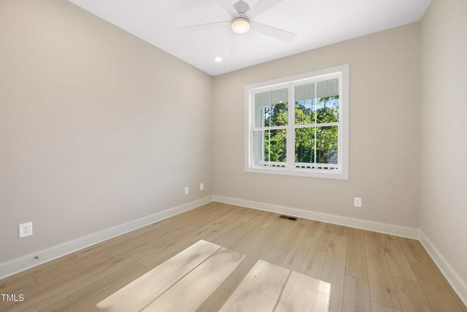 A small, empty room with light wood flooring, beige walls, a white ceiling fan, a window with white trim, and natural light streaming in from outside.