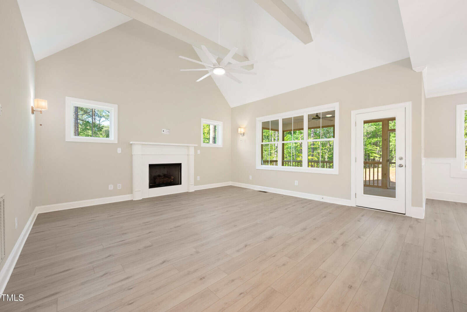 Bright, empty living room with vaulted ceiling, light wood floors, white walls, a ceiling fan, fireplace, and large windows letting in natural light. A glass door leads to an outdoor deck.