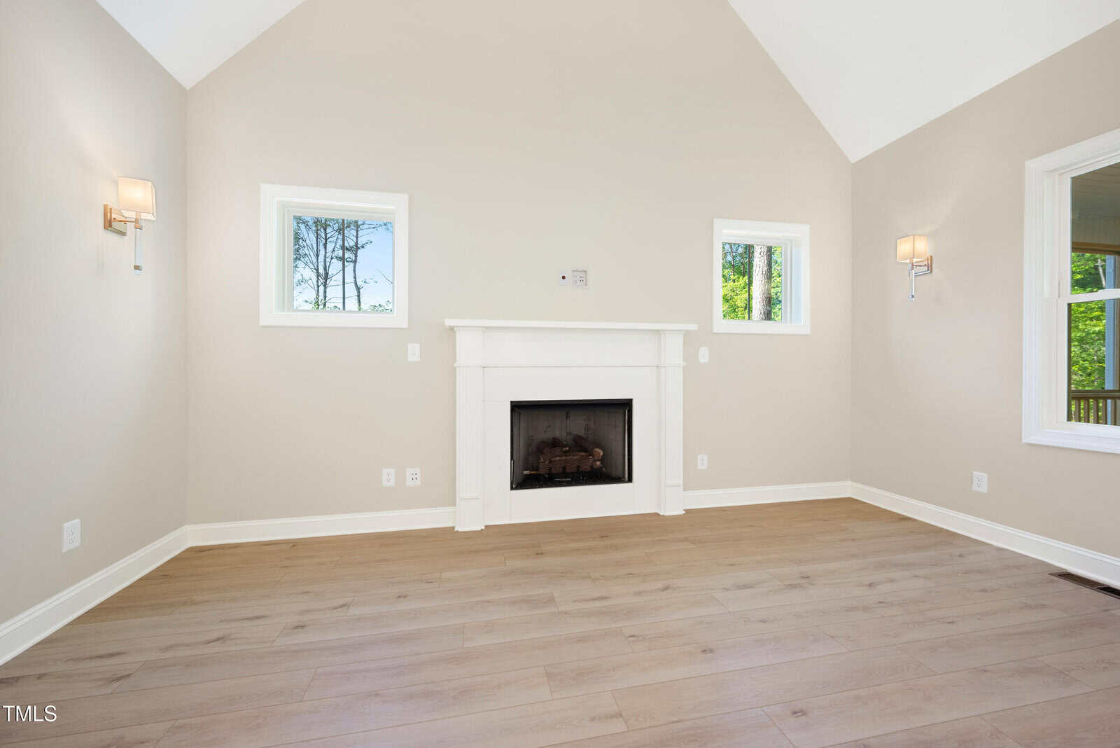 A bright, empty living room with light wood floors, a white fireplace, two wall sconces, and three windows letting in natural light.