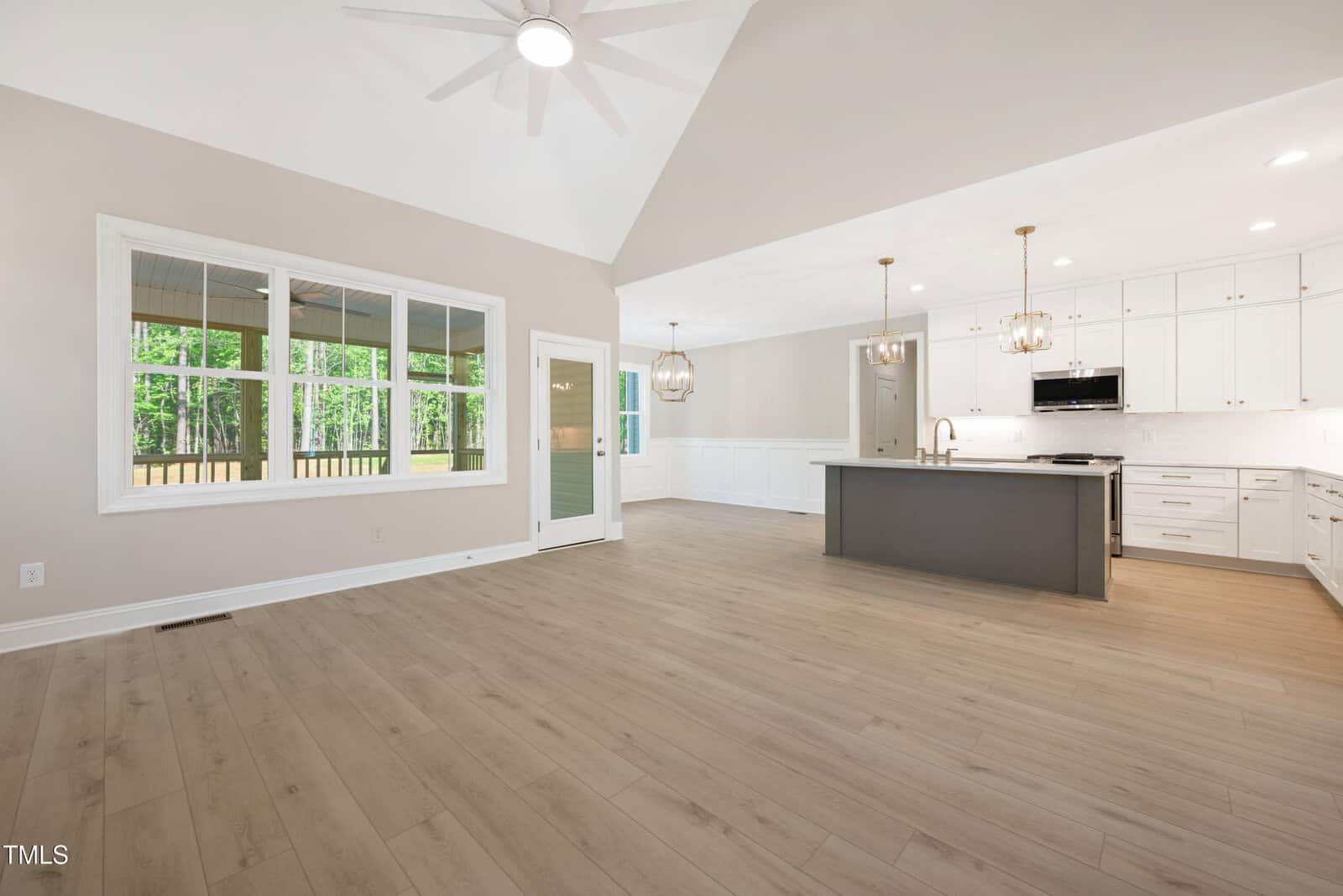 Open-concept living area with light wood flooring, large window, ceiling fan, and modern kitchen featuring white cabinets, a gray island, and pendant lighting. Forest view visible through the windows.
