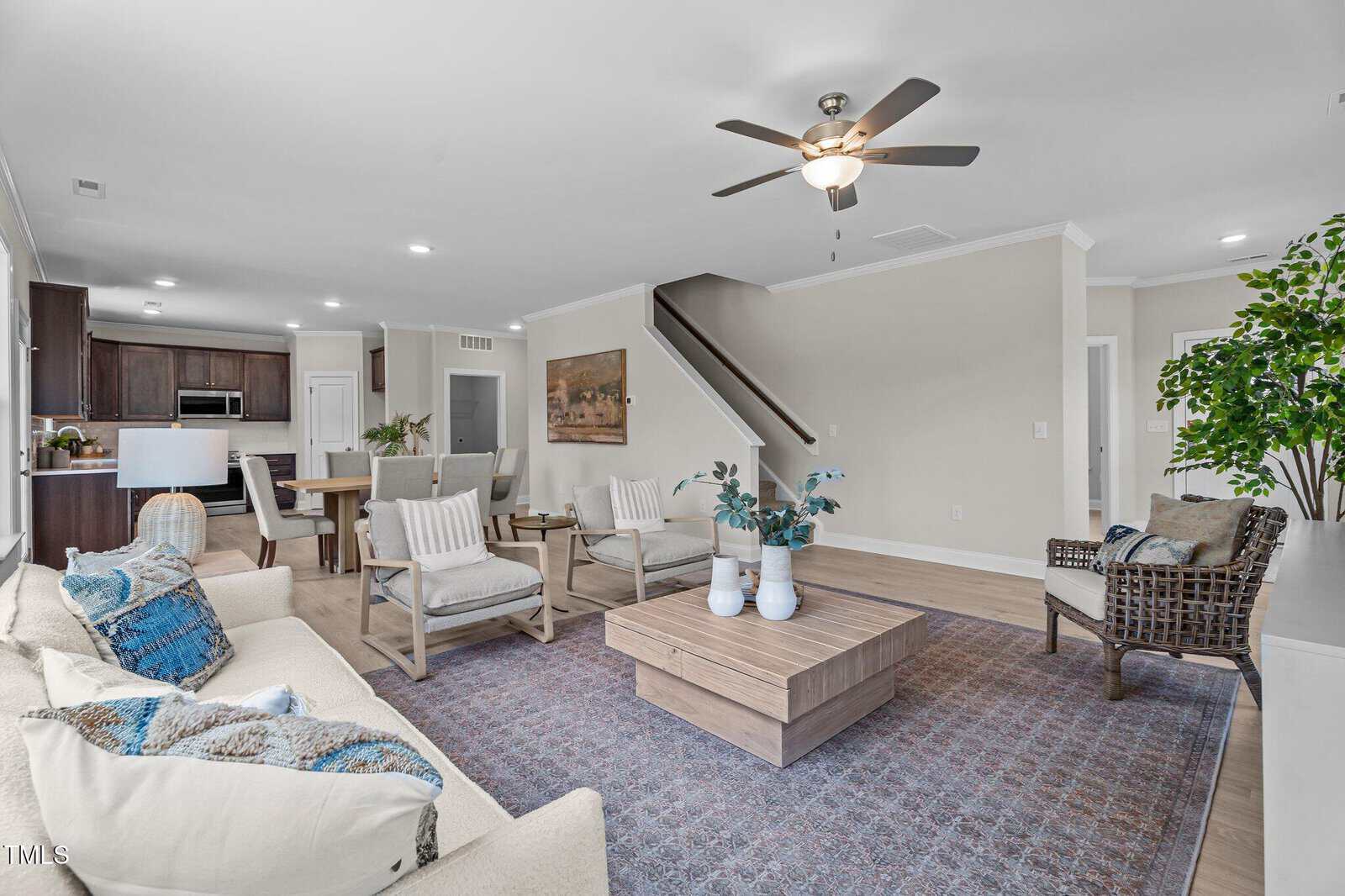 Modern living room with neutral colors, featuring a beige sofa, armchairs, wooden coffee table, large rug, indoor plant, and ceiling fan; kitchen and dining area visible in the background.