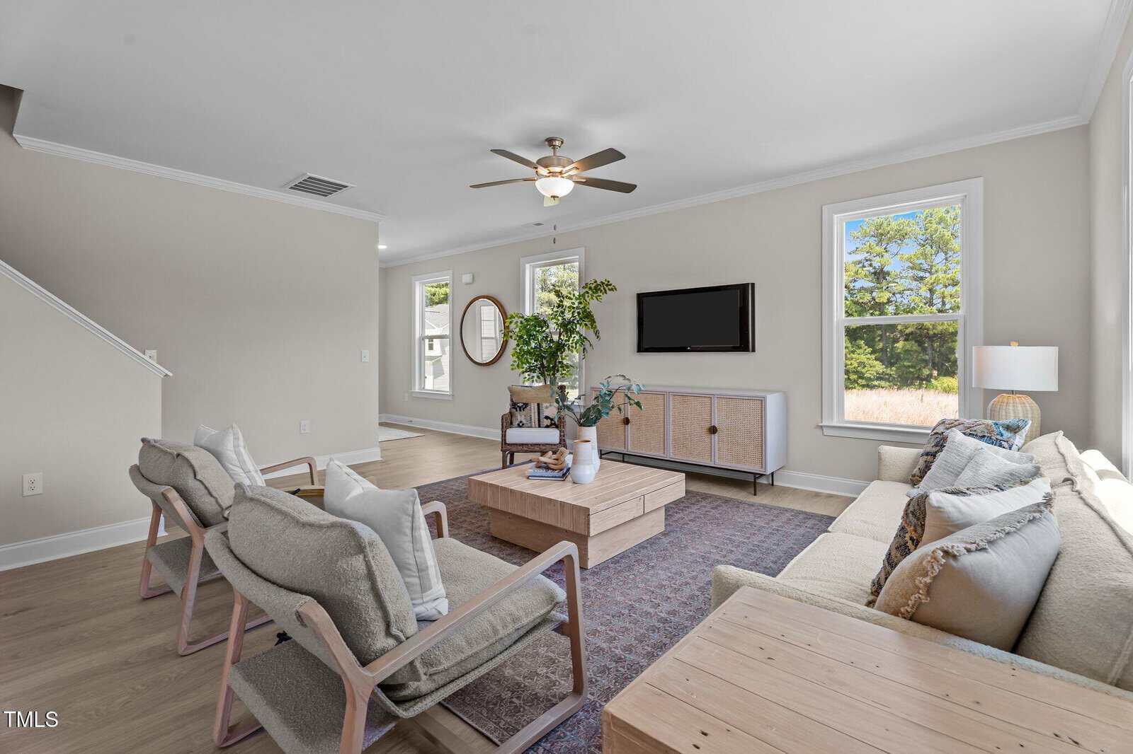 A bright living room with light walls, a ceiling fan, large windows, a beige couch, wooden chairs with cushions, a wooden coffee table, a sideboard, a TV mounted on the wall, and a floor lamp.