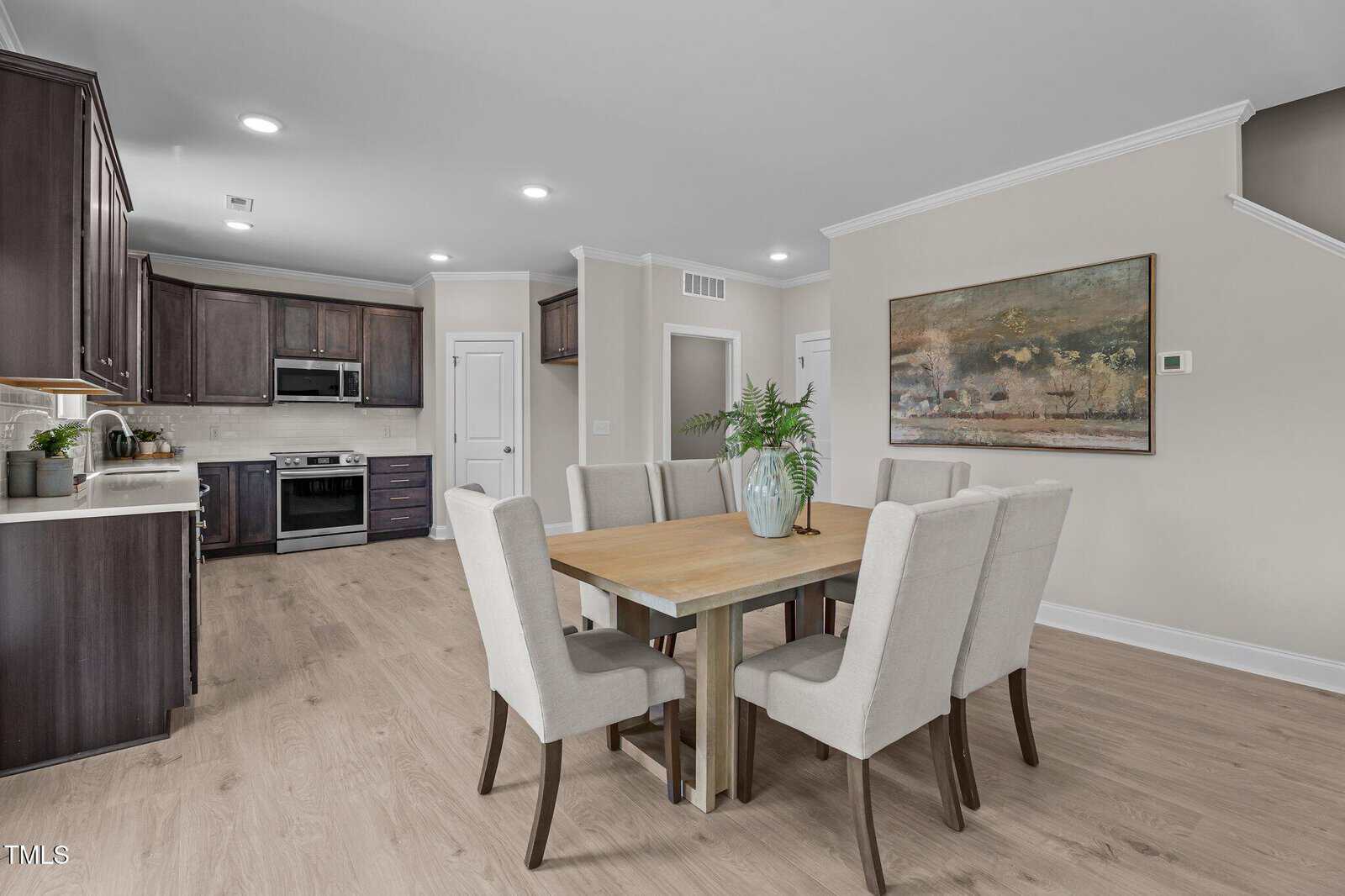 Modern dining area with a wooden table and eight upholstered chairs, adjacent to a kitchen with dark wood cabinets, stainless steel appliances, and light wood flooring. A large abstract painting hangs on the wall.
