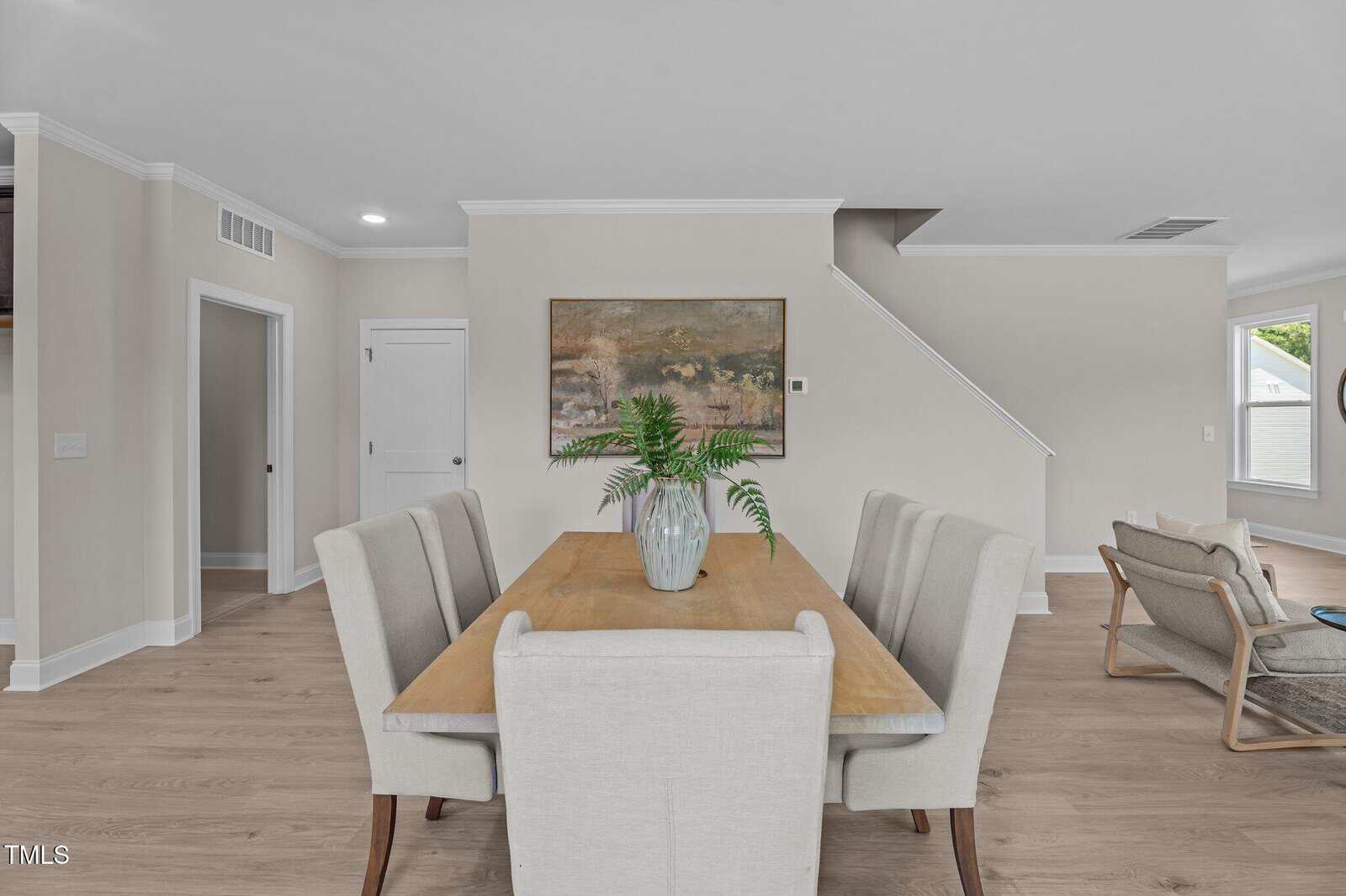 A modern dining area with a wooden table, four beige upholstered chairs, and a glass vase with green ferns. Neutral walls, a staircase, wall art, and part of a living room are visible in the background.