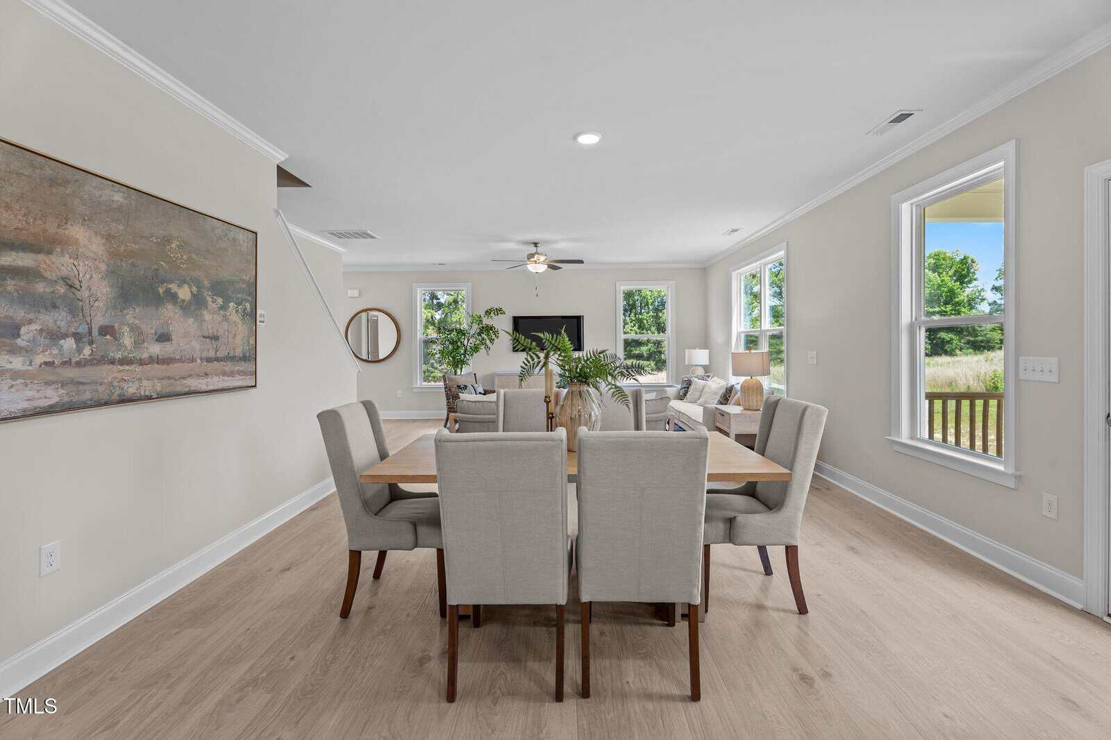 A modern open-concept living and dining area features a wooden dining table with six beige chairs, light wood flooring, neutral walls, large windows with outdoor views, and a cozy living space with sofas and a TV.