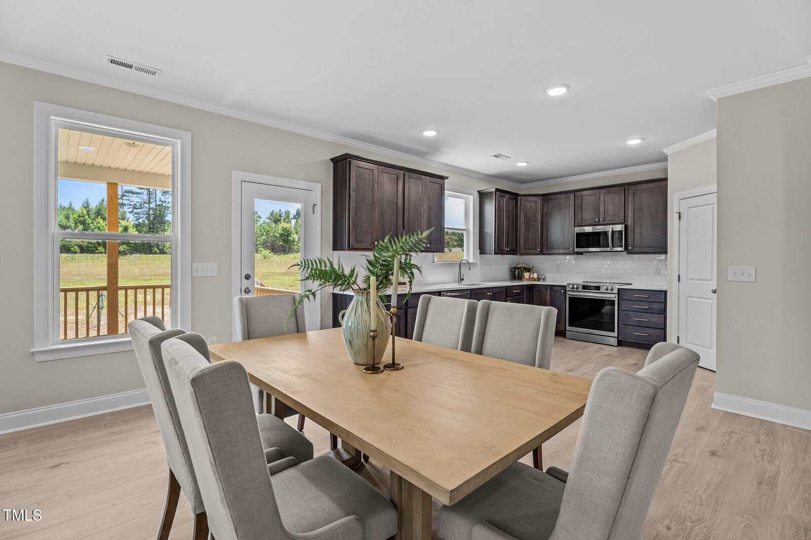 Bright, modern kitchen and dining area with light wood floors, a wooden dining table surrounded by six upholstered chairs, dark cabinets, stainless steel appliances, large windows, and a door to a deck with outdoor views.