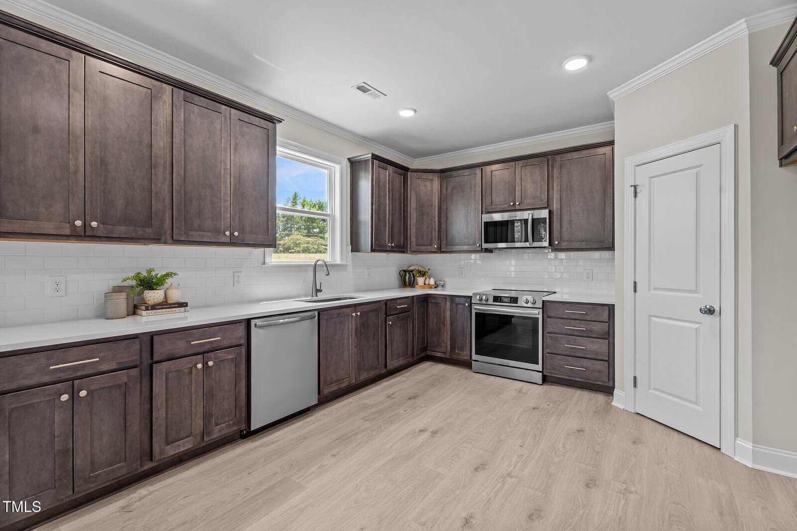 Modern kitchen with dark wood cabinets, stainless steel appliances, light wood flooring, white subway tile backsplash, and a window above the sink letting in natural light.