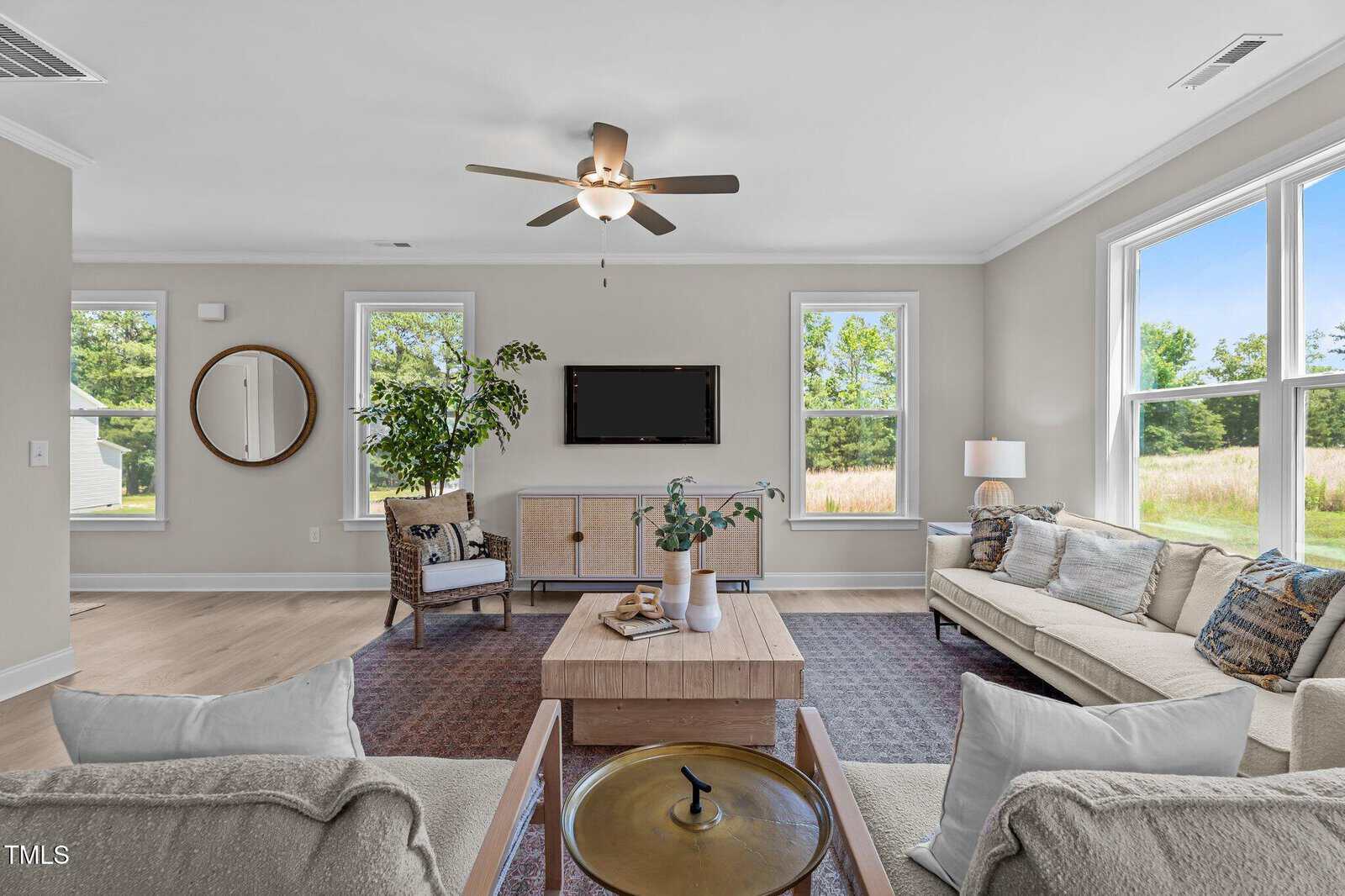Bright living room with large windows, beige walls, and a ceiling fan. Features a light sofa, armchair, coffee table with decor, TV on the wall, and a potted plant near a round mirror. Greenery visible outside.
