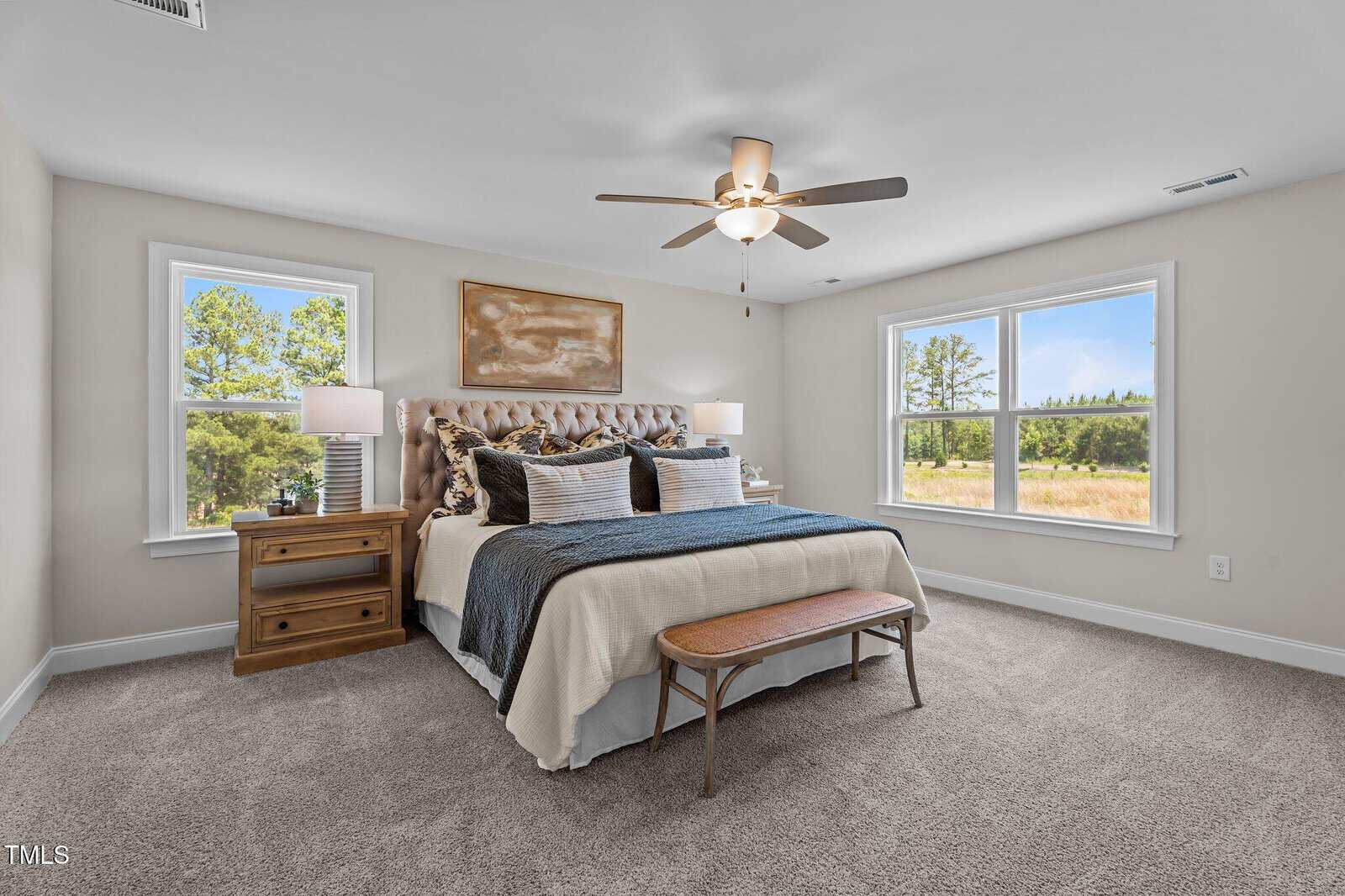 Bright bedroom with a large bed, two wooden nightstands with lamps, a ceiling fan, carpeted floor, bench at the foot of the bed, and two large windows showing a scenic outdoor view.