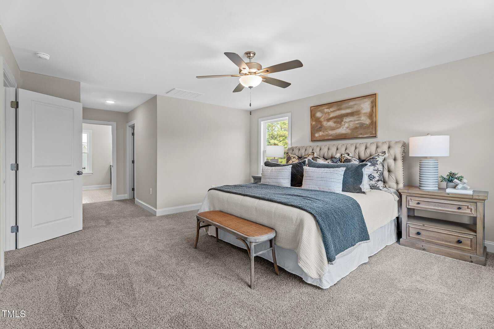 A spacious bedroom with a large bed, blue and white bedding, a wooden bench at the foot, nightstand with lamp and decor, ceiling fan, beige walls, carpeted floor, and open doors to hallway and another room.
