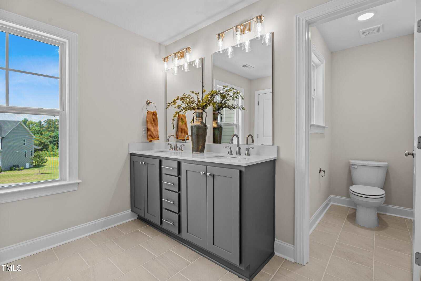 Bright bathroom with double-sink vanity, dark gray cabinets, large mirror, two light fixtures, and decorative vases. Toilet is in a separate nook, window shows green yard outside. Neutral tile flooring throughout.
