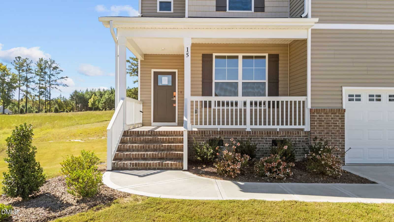 A tan house with a brick foundation, white railings, and brown shutters. A short set of stairs leads to a covered front porch. Landscaping with bushes lines the sidewalk, and grass extends into a field beyond.
