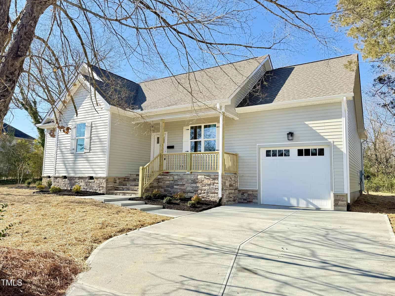 A yellow single-story house with a steep roof, attached garage, and a small front porch. The front yard has newly planted shrubs and a concrete driveway. Leafless trees are visible around the property.