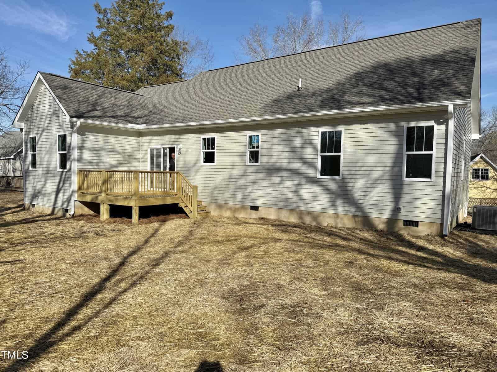 A single-story house with light-colored siding, multiple windows, and a small wooden deck in the back. The yard is covered with straw, indicating recent landscaping work. The sky is clear with some trees casting shadows on the house.