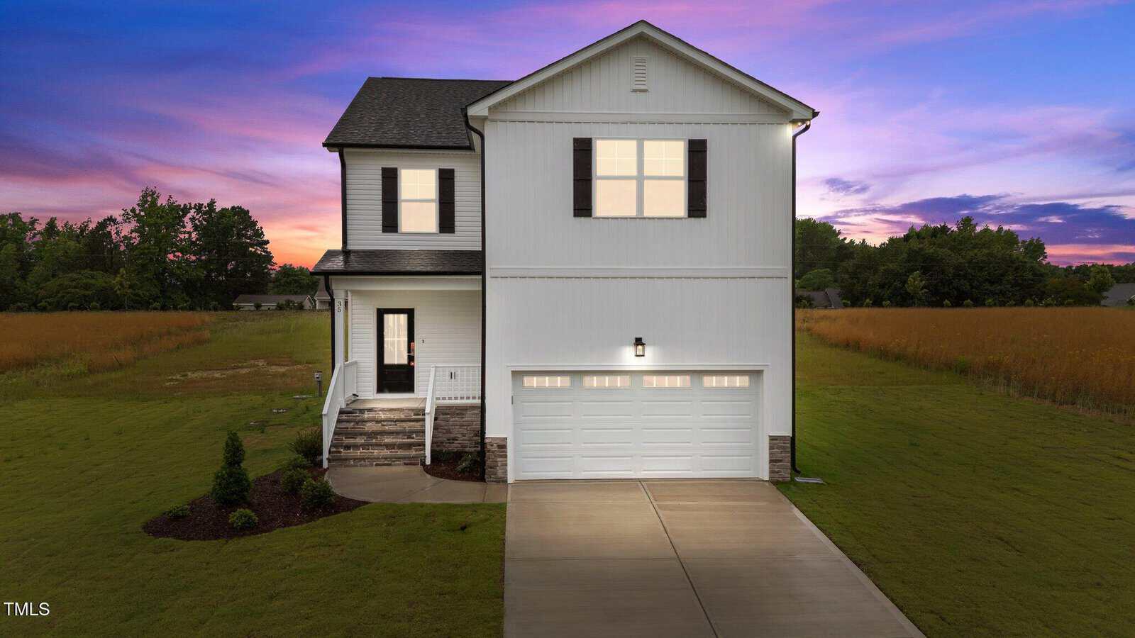 Two-story white house with black shutters and a double garage, set on a large grassy lot with a paved driveway. The sky is purple and pink at sunset, and there are trees in the background.