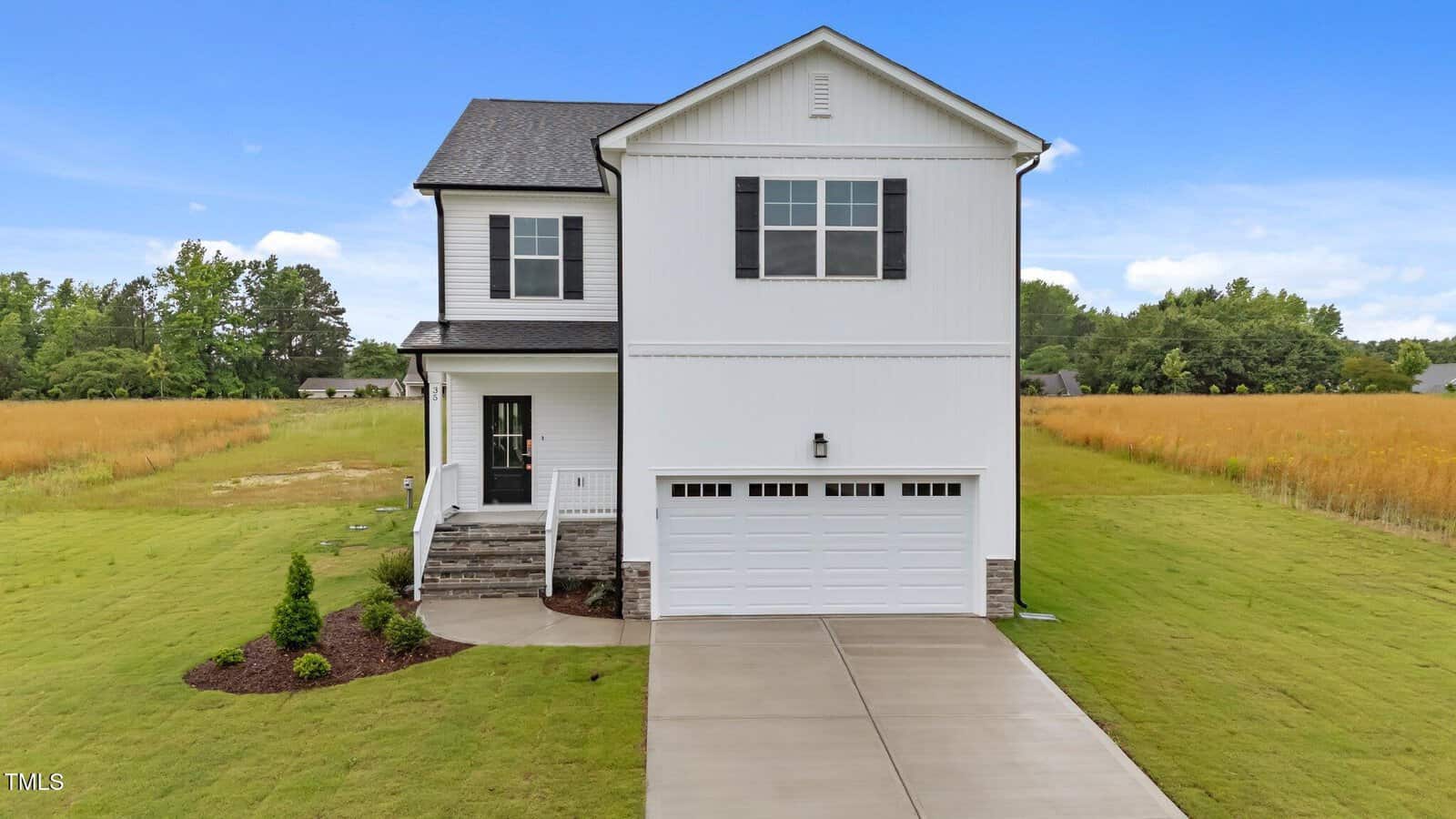 A two-story white house with black trim, a covered front porch, and a double garage. The home is surrounded by a grassy yard and fields, with a concrete driveway leading to the garage.