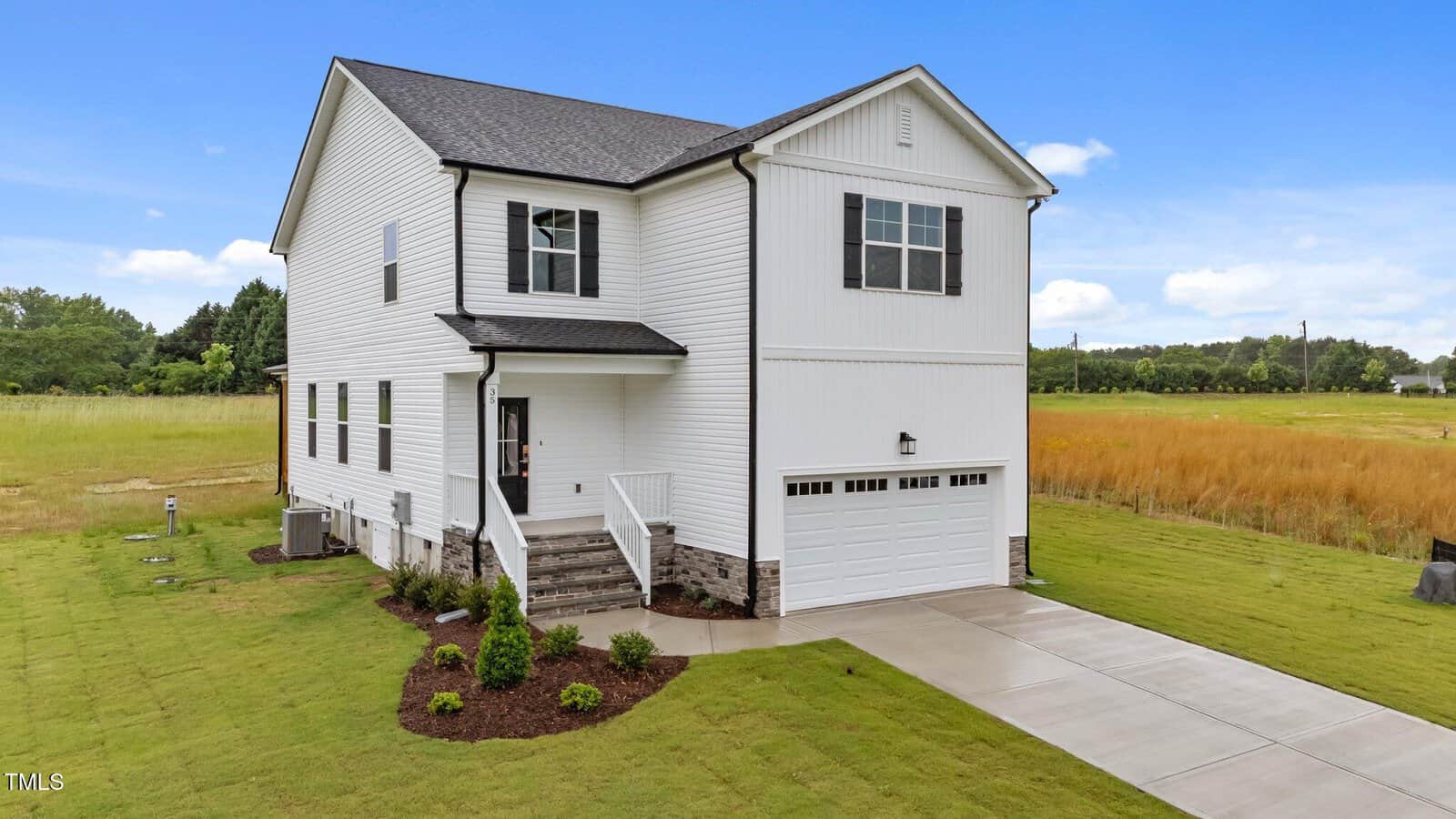 Two-story white house with black roof, attached two-car garage, and small front porch, set on a large grassy lot with a concrete driveway and minimal landscaping, under a partly cloudy sky.