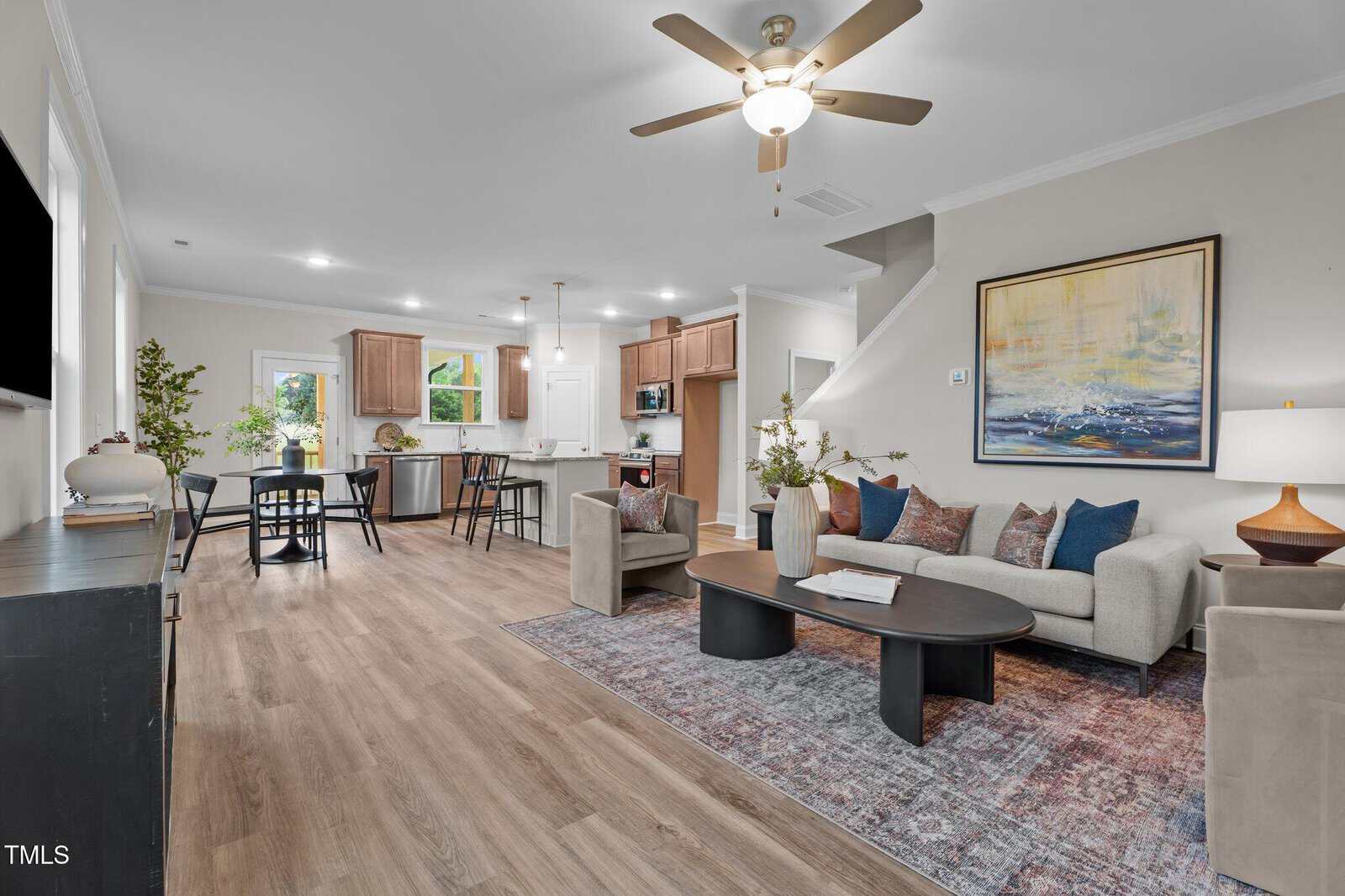 Modern open-concept living room and kitchen with wood floors, neutral tones, a ceiling fan, large painting, dining table, and stylish furniture. Natural light streams in from windows and a glass door.