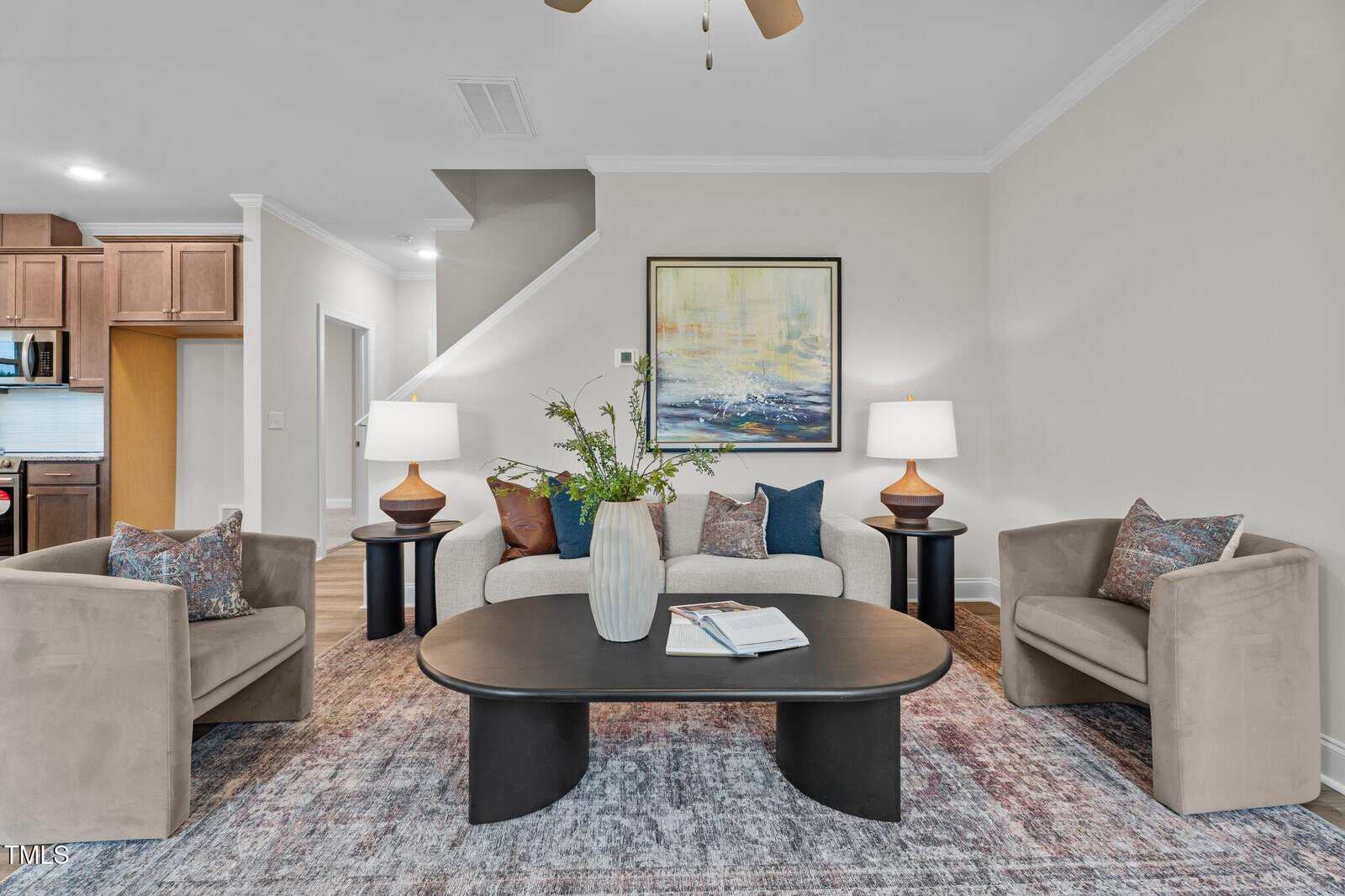 A modern living room with a beige sofa, two armchairs, a black oval coffee table, a large vase with greenery, an open book, two table lamps, a framed painting, and a staircase in the background.