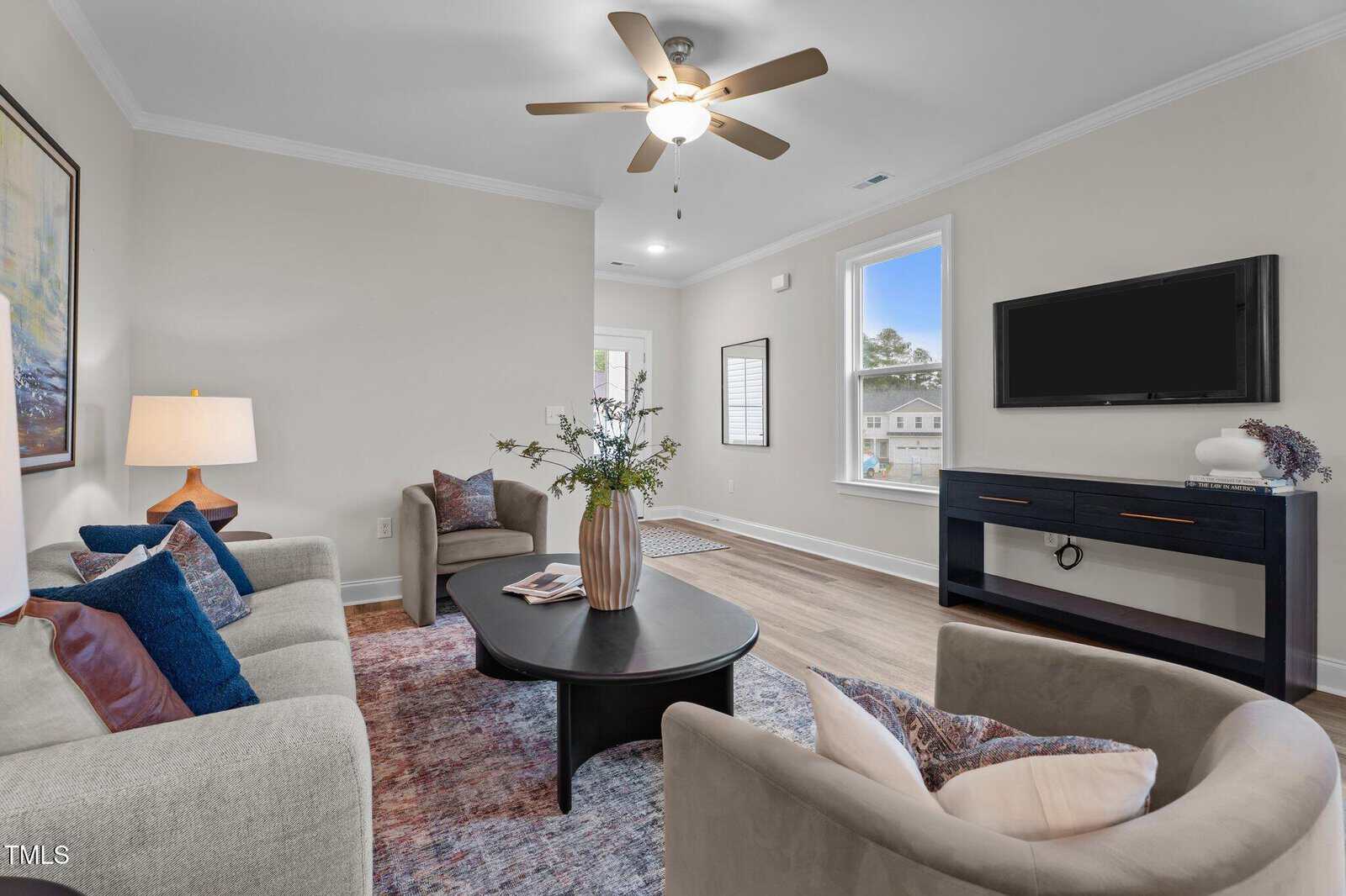 A modern living room with two beige armchairs, a beige sofa, a black coffee table with a vase, a rug, a ceiling fan, a floor lamp, wall art, a flat-screen TV on a black console, and large windows.