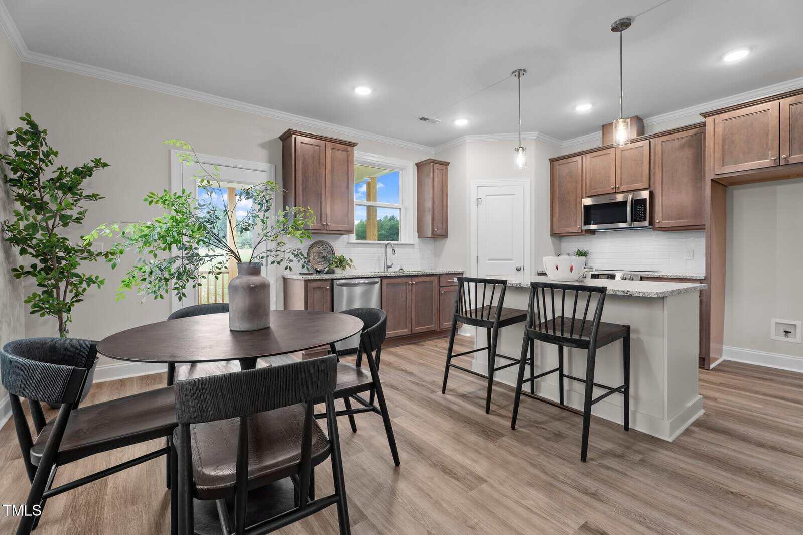 Modern kitchen and dining area with wood cabinets, white countertops, stainless steel appliances, a round dining table with four chairs, barstools at the island, plants, and light wood flooring.