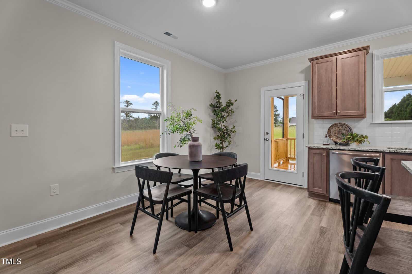 A dining area with a round black table and four black chairs sits next to a window. There are plants, wood cabinets, and a glass door leading to a porch, with a view of grassy fields outside.