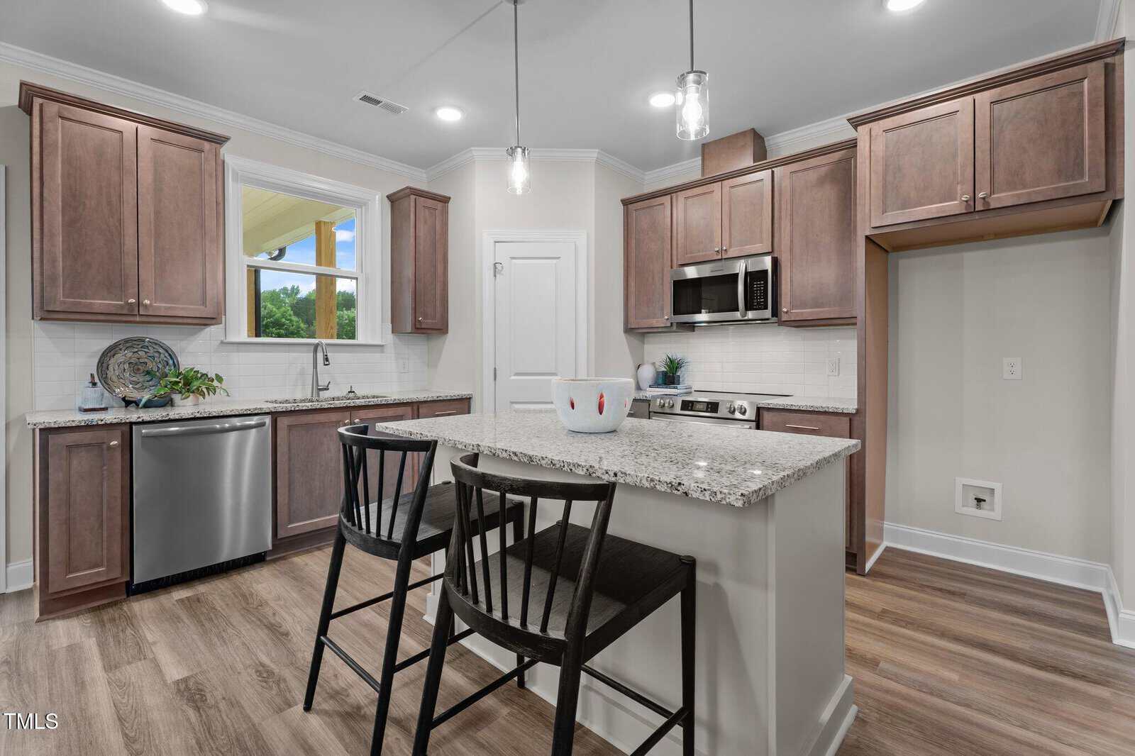 Modern kitchen with brown cabinets, stainless steel appliances, and an island with two black chairs. Granite countertops, a dishwasher under the window, and wood-style flooring are visible. The space appears clean and well-lit.
