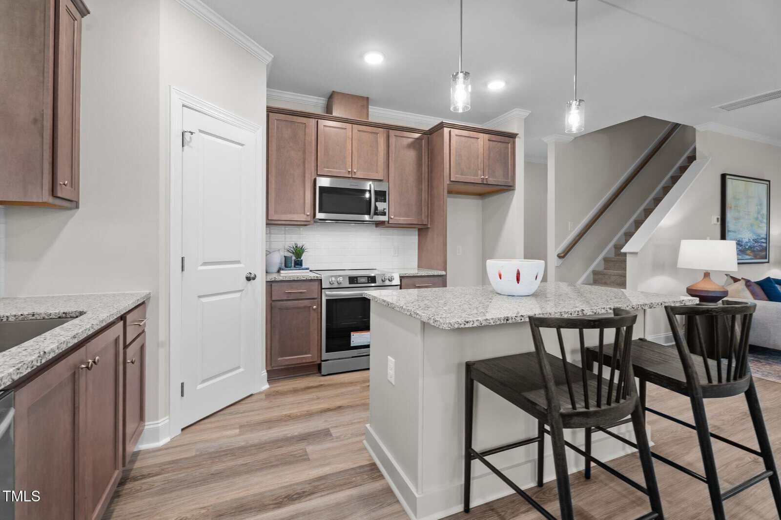 A modern kitchen with brown wooden cabinets, stainless steel appliances, and a granite island with two black chairs. Pendant lights hang above the island, and stairs lead to the upper floor in the background.