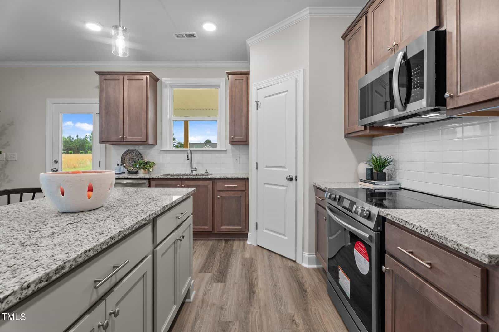 Modern kitchen with brown and gray cabinets, granite countertops, stainless steel appliances, an island with a white bowl, wood-style floor, and a window above the sink showing a view of a field outside.