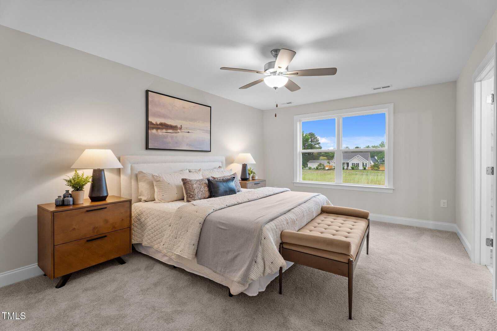 Bright bedroom with a king bed, beige carpet, nightstands with lamps, art above the headboard, a bench at the foot of the bed, and a window showing a green lawn and other houses.