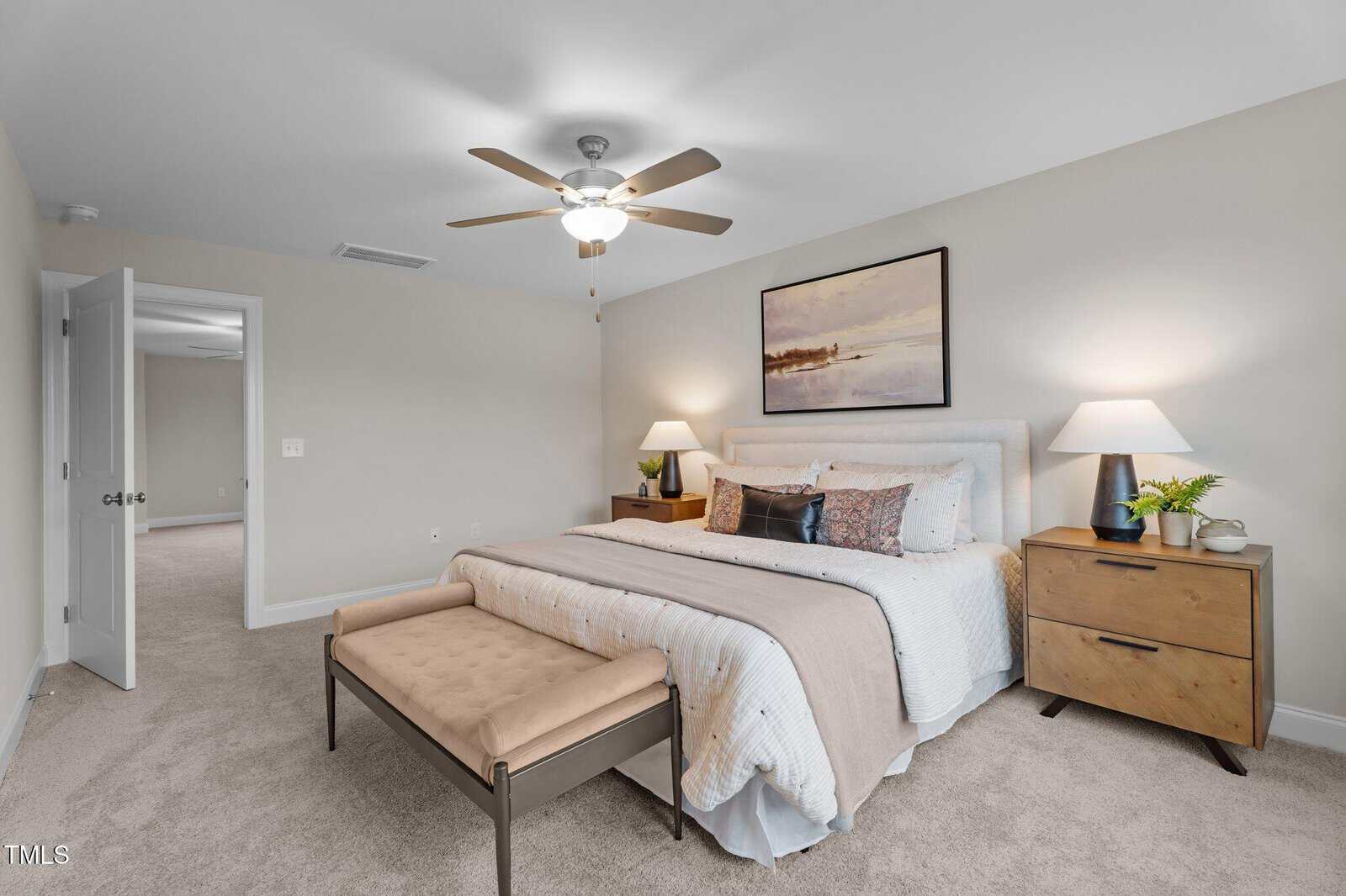 A neatly made bed with neutral bedding, two wooden nightstands with lamps, a bench at the foot, and a nature painting above the headboard in a bright, carpeted bedroom.
