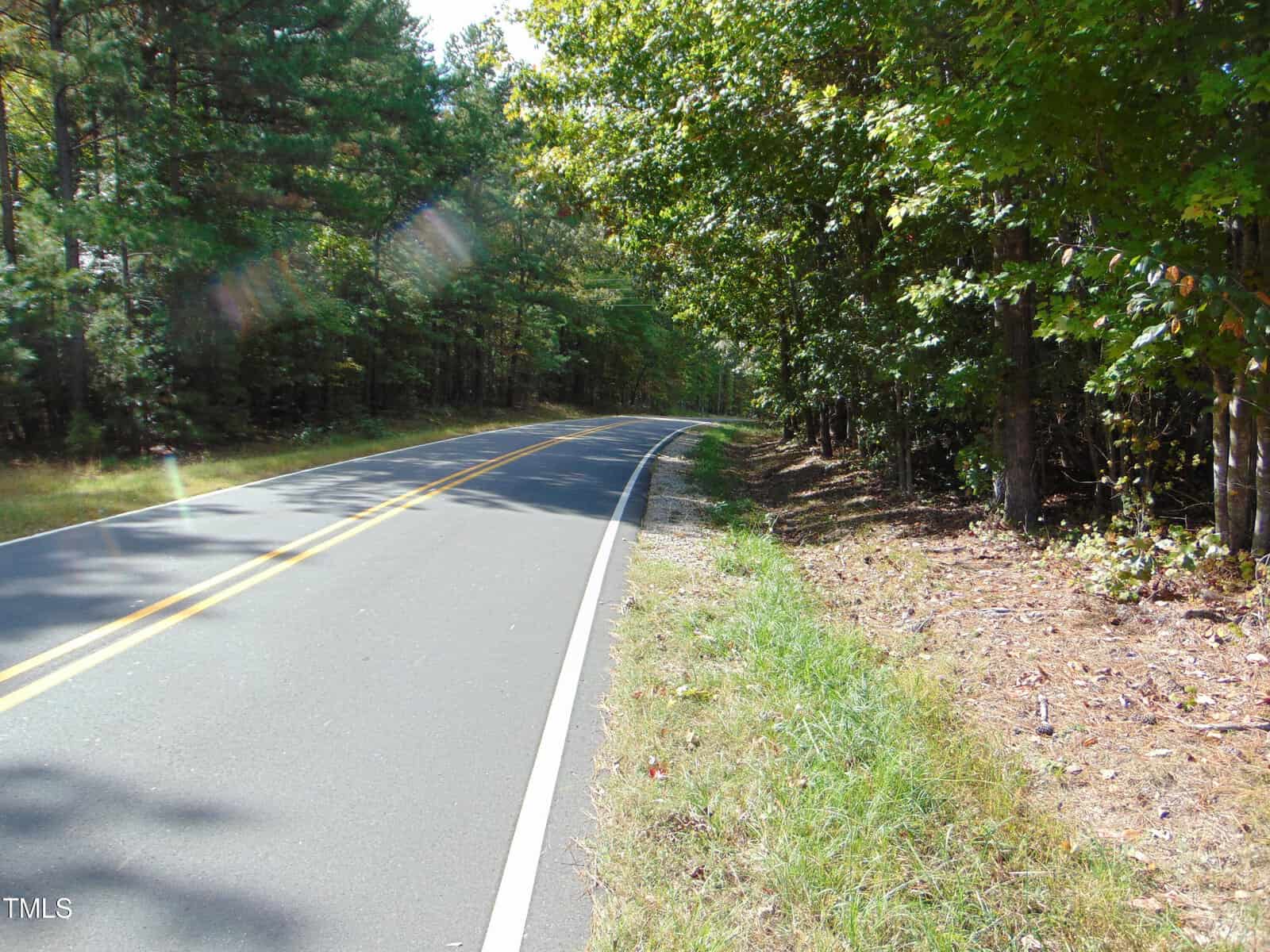A winding road named Bullock flanked by dense green forest under a partly cloudy sky, with sun rays filtering through the trees. The road, reminiscent of 0 Tungsten Mine Road, features a yellow center line and white edges, suggesting a peaceful rural or suburban setting.