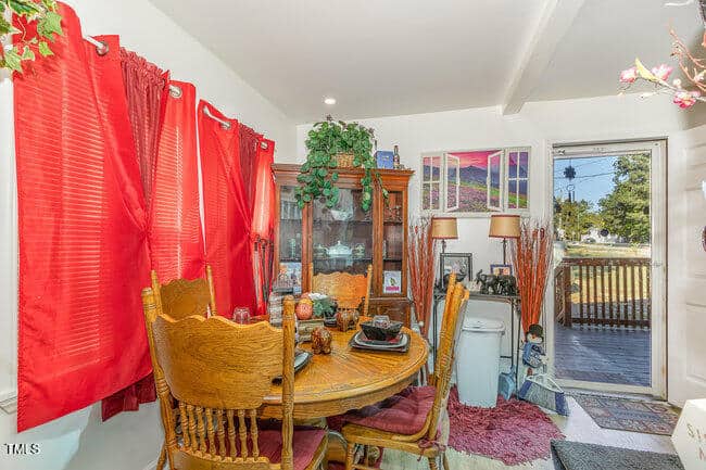 A cozy dining area with a wooden table and chairs, decorated with red curtains and a china cabinet filled with dishes. Plants and photos adorn the room, and a door opens to a porch with a view of trees and power lines outside.