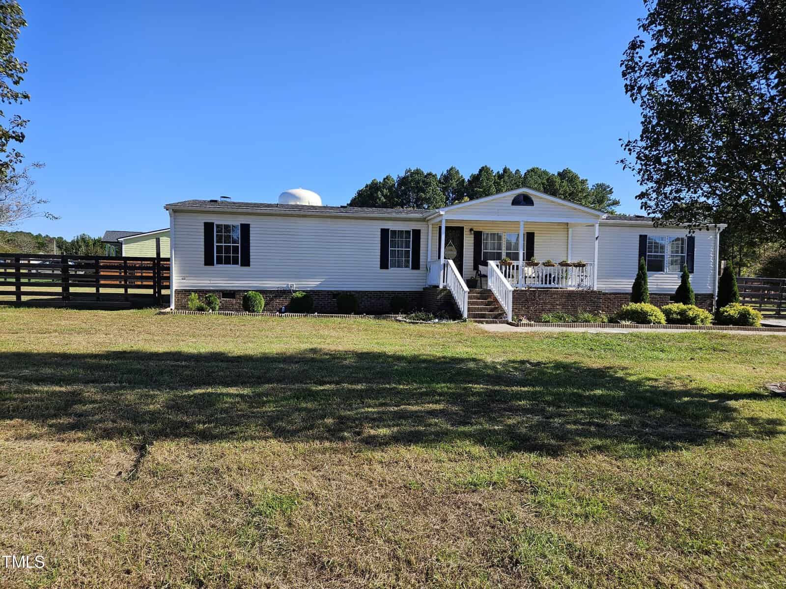 The single-story white house at 2084 Meadow View Drive boasts a charming porch, surrounded by a well-maintained lawn and trees in the heart of Creedmoor. A wooden fence lines the property, all under a clear blue sky.