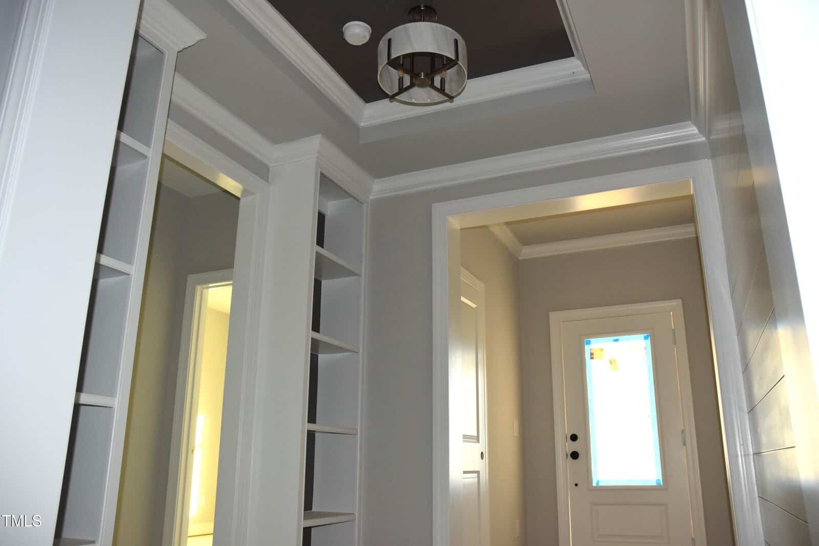A hallway with light gray walls and white trim features built-in shelves on the left, a white door at the end, and a ceiling light fixture. A transom window above the door lets in natural light.