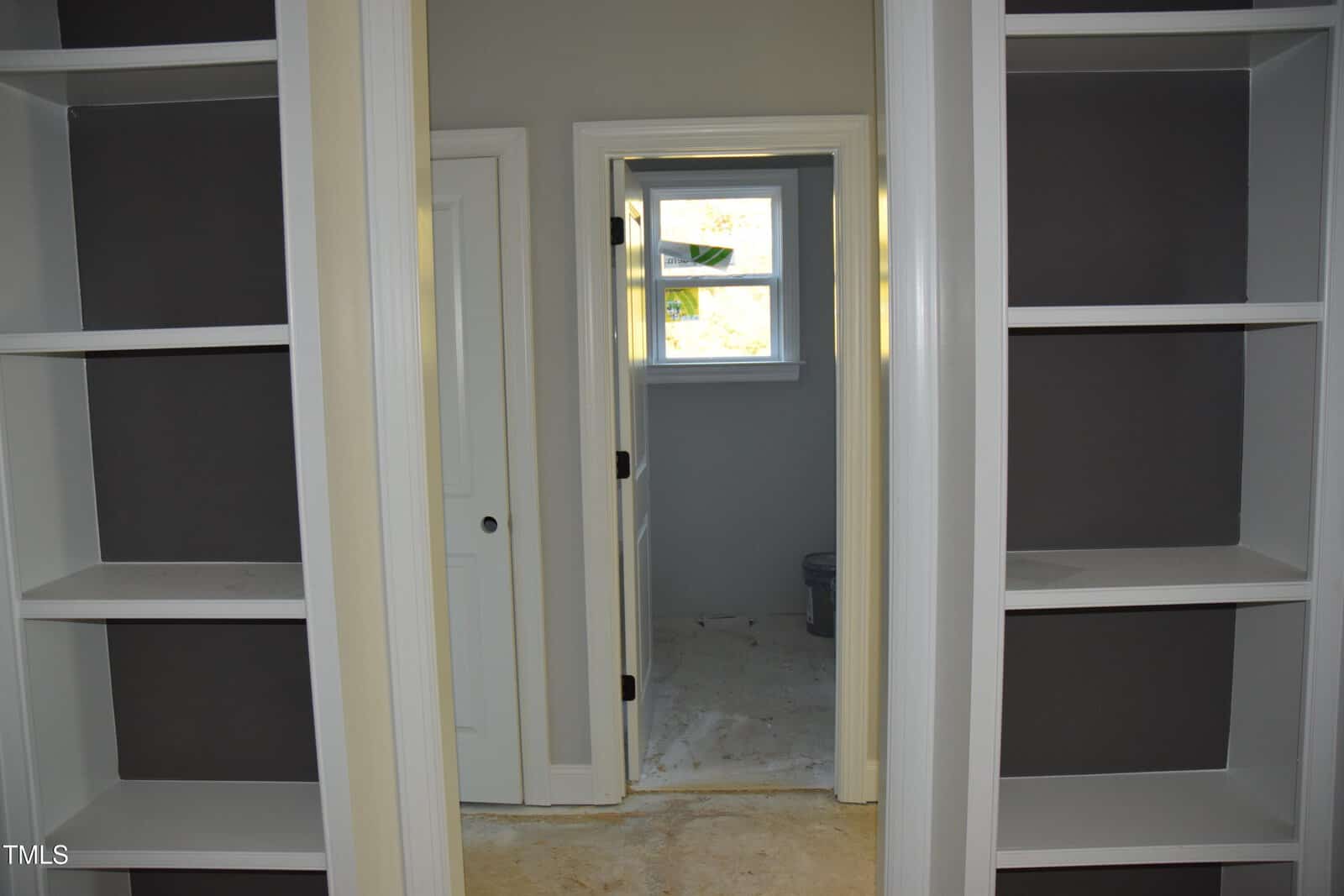 The image shows an unfinished interior with two empty white built-in bookshelves on either side of a doorway. Beyond the doorway is another room with a window and a toilet visible, with unfinished flooring throughout.