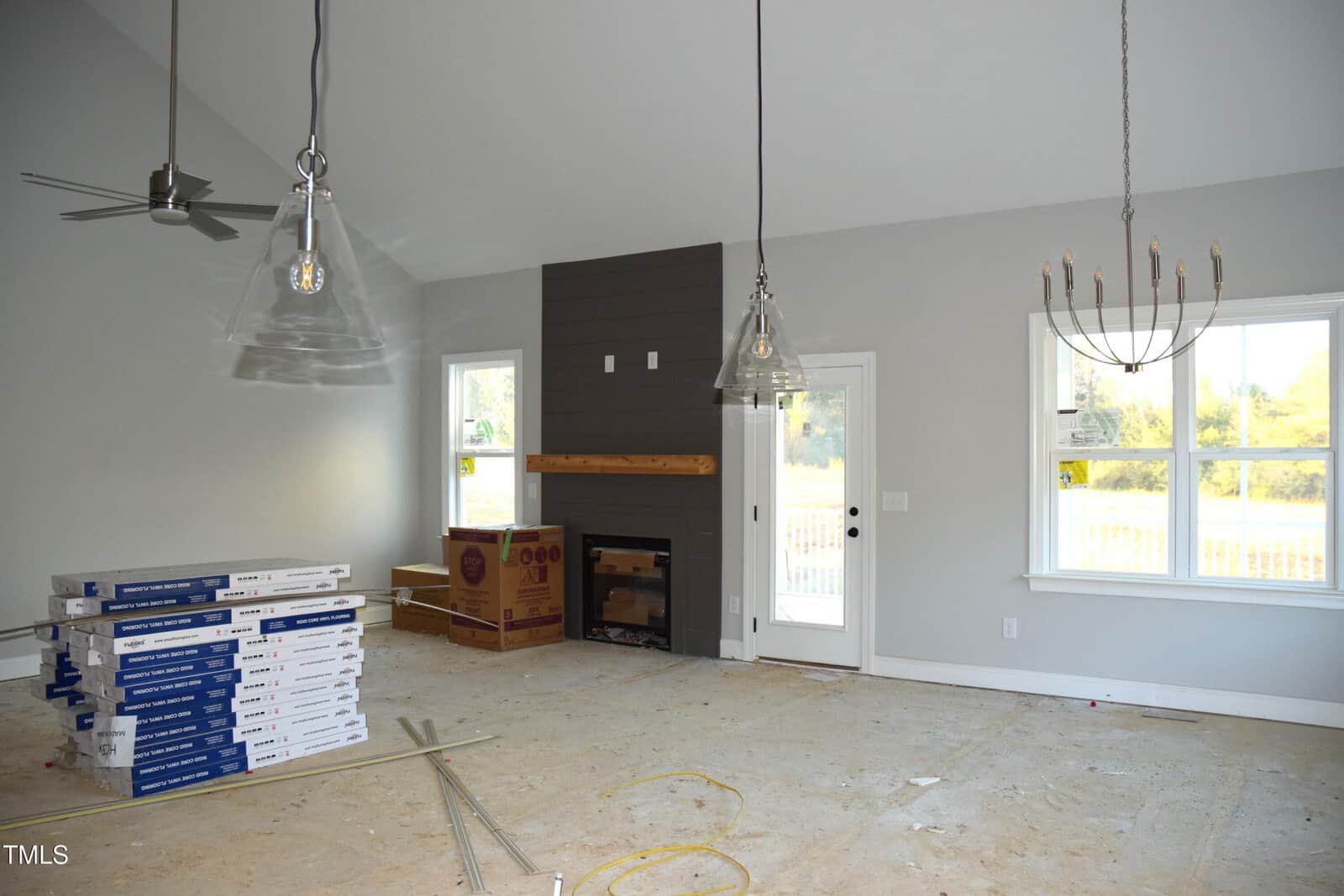 A partially constructed living room with a vaulted ceiling, featuring a dark accent wall with a fireplace, boxed light fixtures on the floor, and stacked wooden planks. Large windows and a glass door let in natural light.