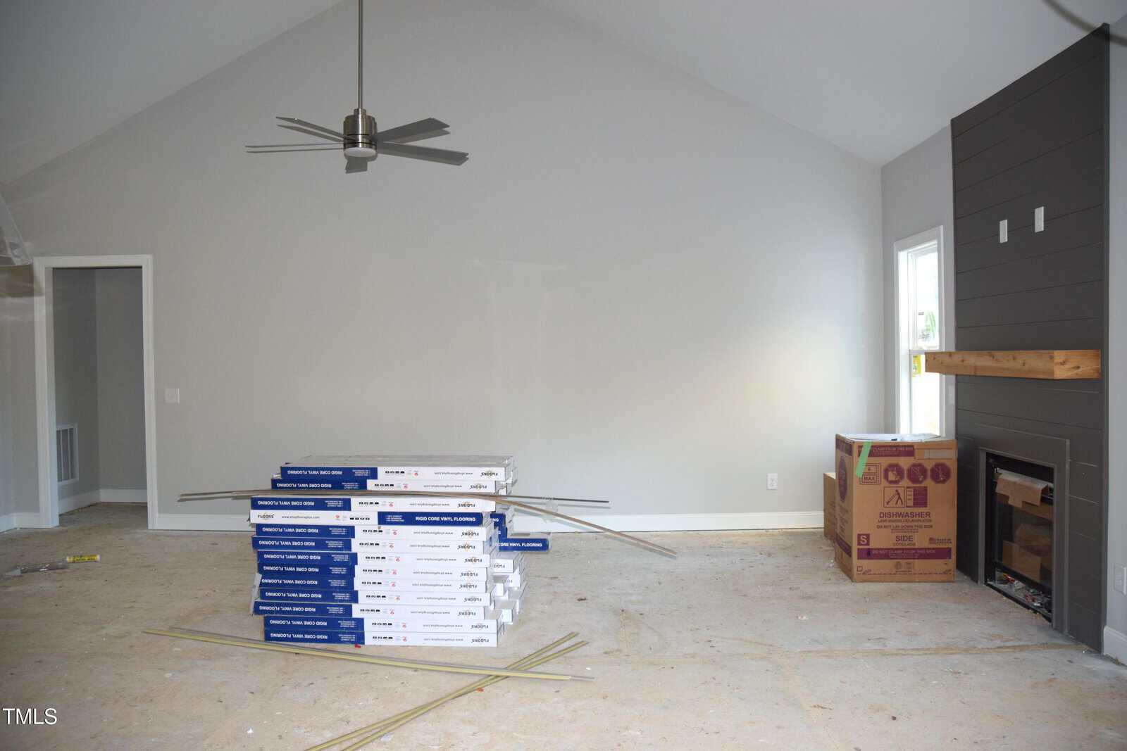A room with unpainted walls and bare floors. Flooring materials are stacked in the center, along with a cardboard box. A ceiling fan is installed, and there is a fireplace with a wooden mantle on the right. A window brings in natural light.