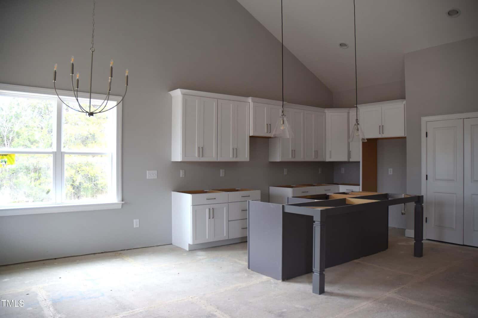 A modern kitchen under construction with unpainted concrete floors. It features white cabinets, a gray island without a countertop, and pendant lights. A large window on the left side lets in natural light.