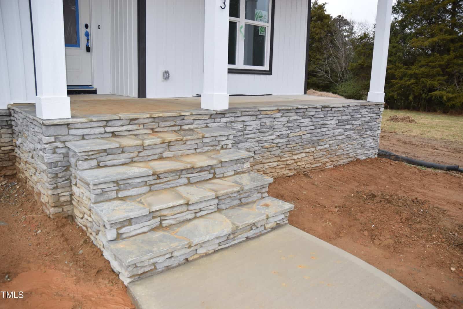 Stone steps and a path lead to a white-sided building's entrance. The porch area is elevated, featuring a stone facade. The ground around the path is bare, with some grass and trees in the background.