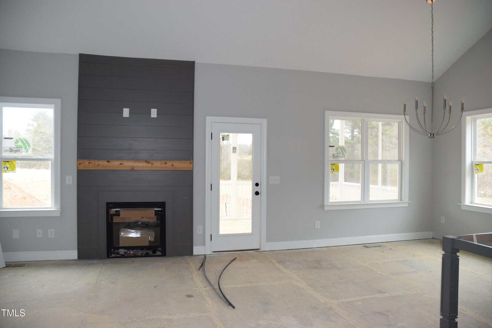 A room with light gray walls and a vaulted ceiling. It features a dark gray paneled fireplace with a wooden mantel, two windows, a glass door, and an unlit chandelier. The floor is bare and appears to be undergoing renovation.