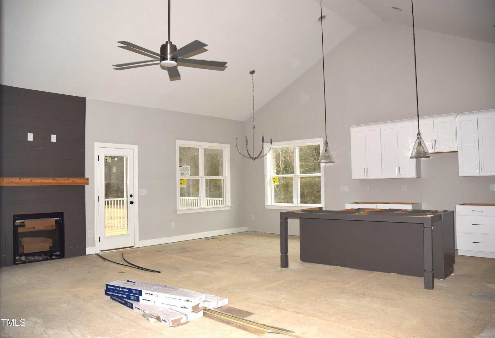 Spacious, unfurnished kitchen and living area with high ceilings. Features include a modern ceiling fan, large windows, a fireplace, white cabinetry, and a center island. The floor is unfinished, with building materials visible on the concrete.