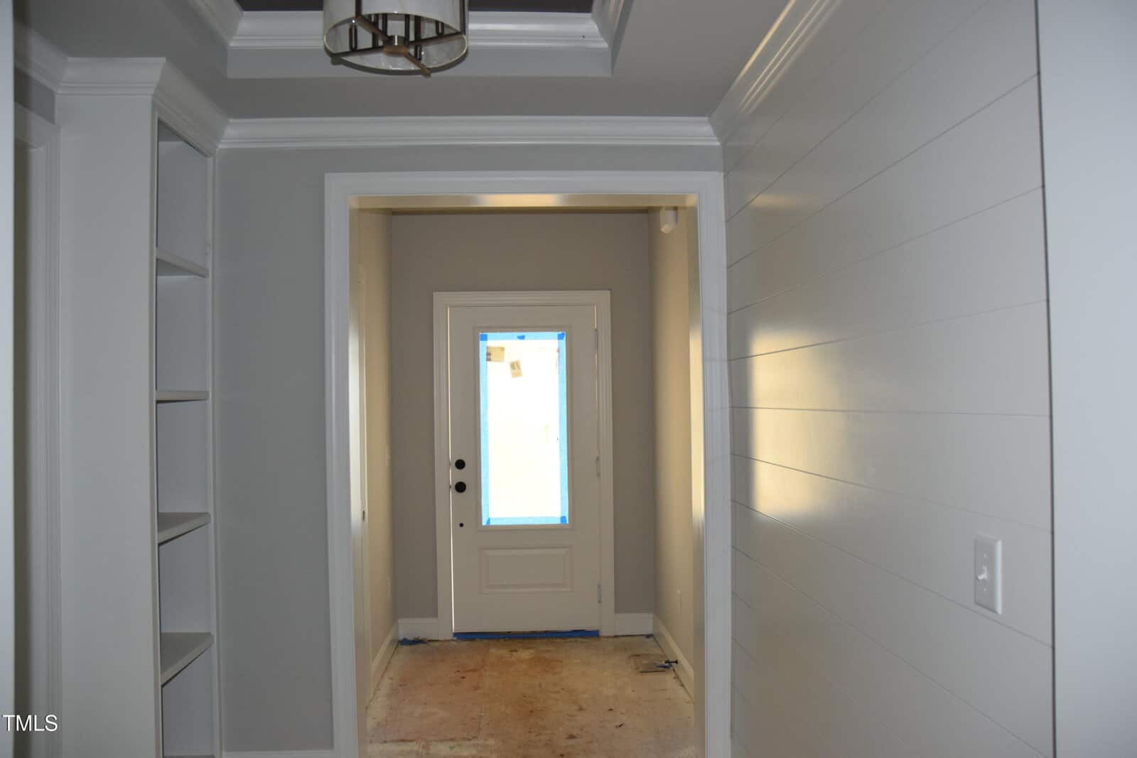 A hallway with light gray walls and ceiling leads to a white front door with a window. The floor is unfinished, and there is a built-in shelving unit on the left. A ceiling light fixture hangs above. The space is well-lit by natural light.