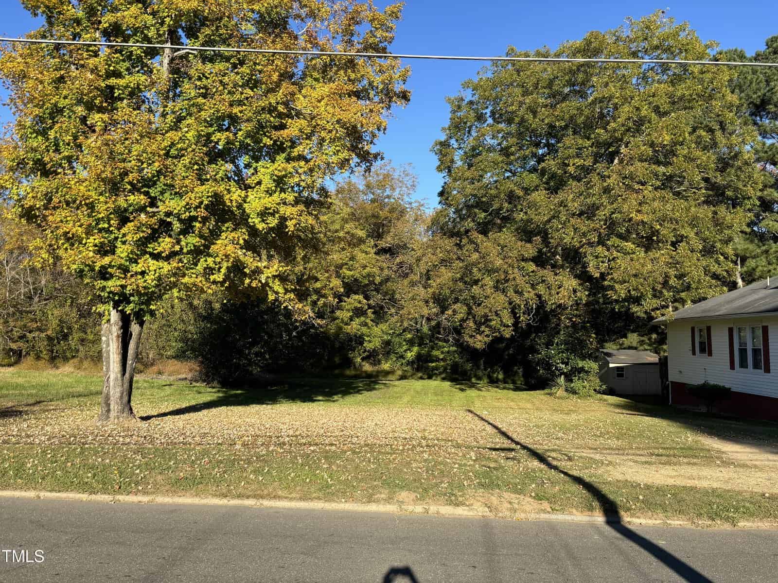A quiet suburban street features a large, grassy yard bordered by tall trees with autumn foliage on the left. A small white house with a red base sits on the right. The sky is clear and blue.