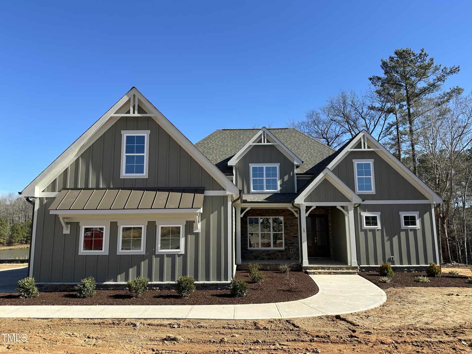 A newly constructed two-story house with gray board and batten siding, white trim, a brown shingle roof, a covered front porch, and landscaped shrubs along a curved concrete walkway.