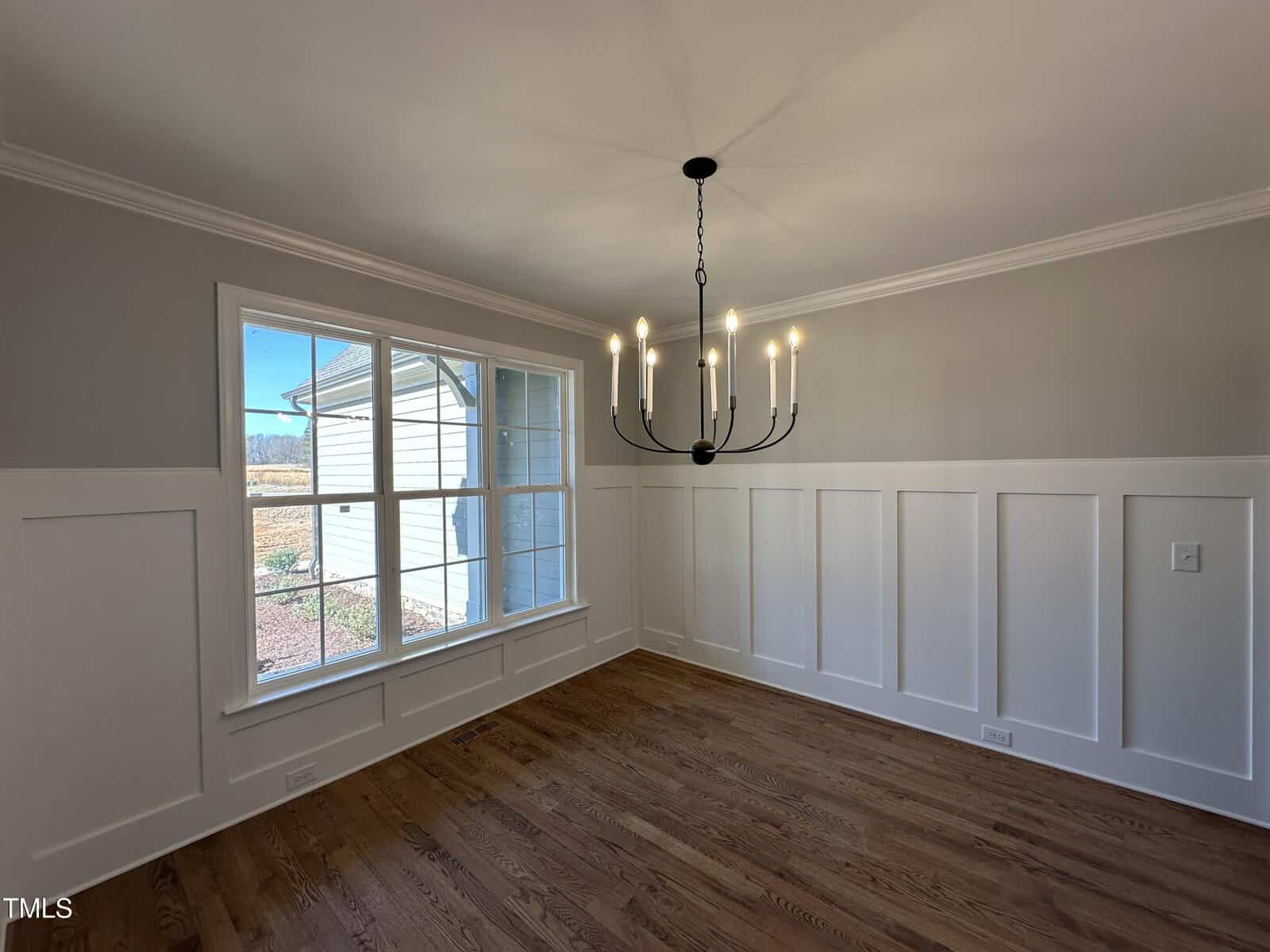 A bright, empty room with hardwood floors, white wainscoting, beige upper walls, a large window, and a modern black chandelier hanging from the ceiling. Natural light enters through the window.