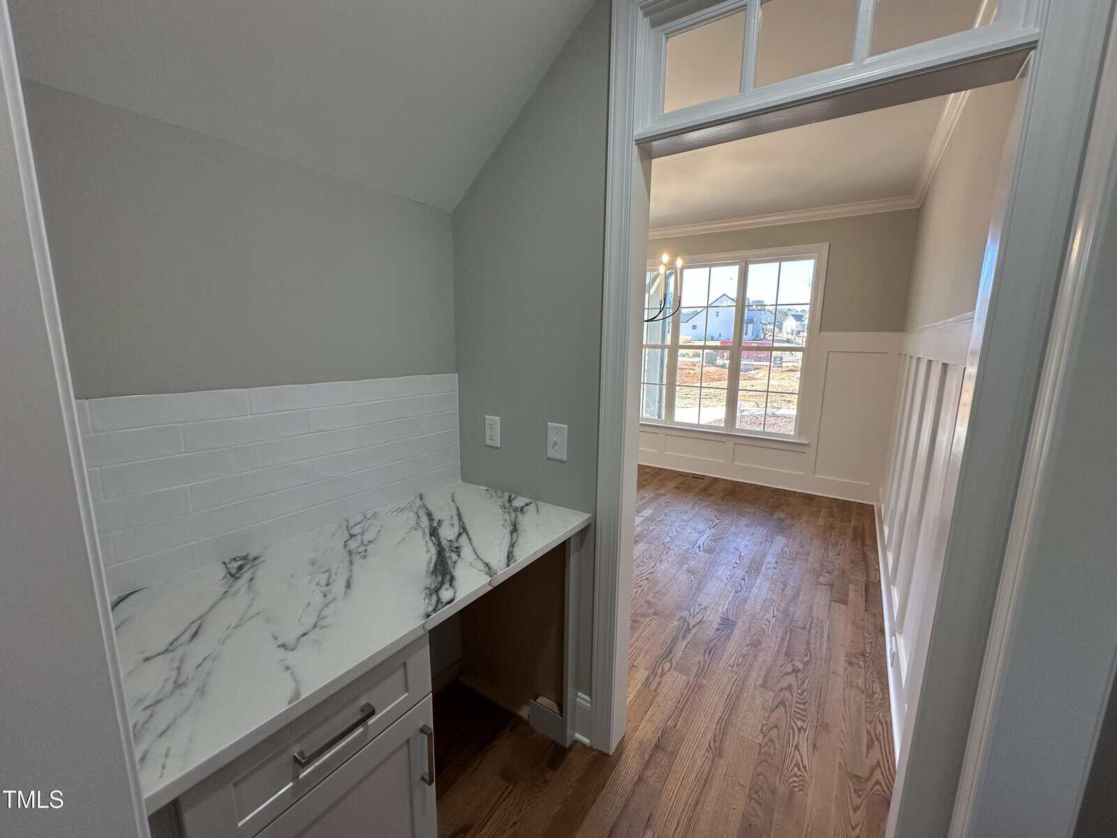A small built-in desk area with a marble-patterned countertop, white tile backsplash, and drawers, opens into a bright room with hardwood floors, wainscoting, large windows, and views of a neighborhood outside.